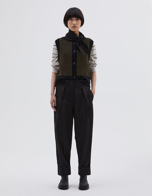 Full body image, front view of model wearing black cotton wool trousers, with zip fly and corozo brace button fastening, self waistband detail, slant pockets, front pleats and deep stitch hem. Styled with white and black striped long sleeve top, knitted green waistband with black trim, black knitted scout scarf and black leather boots.