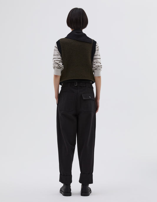Full body image, back view of model wearing black cotton wool trousers with self waistband with cinch back and belt loops, button through flap patch and patch back pockets. Styled with white and black striped long sleeve top, knitted green waistband with black trim, black knitted scout scarf and black leather boots.