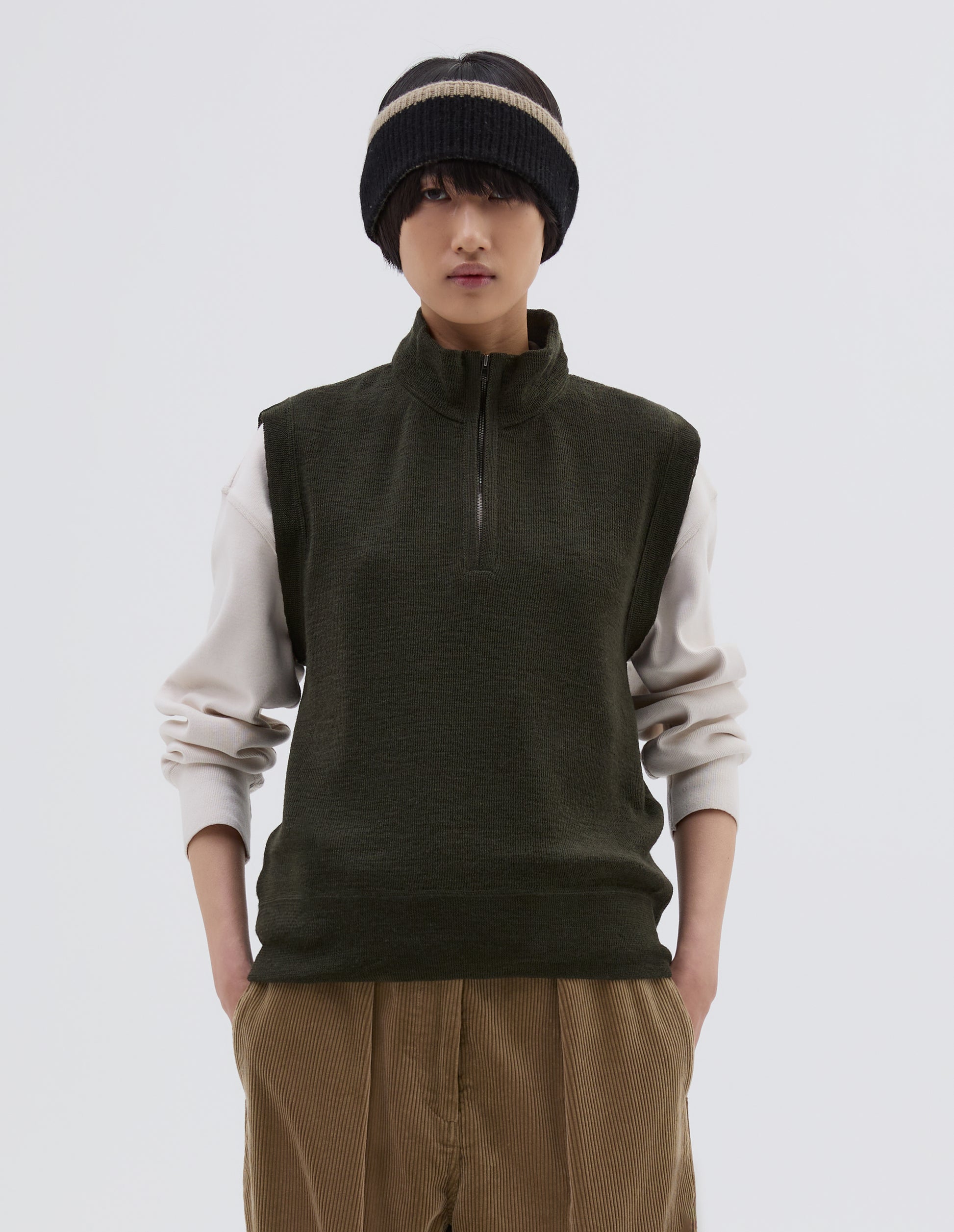 Cropped body image, front view of model wearing dark green wool slipover, with half zip mock turtleneck and flatlock stitch detailing on seams. Styled over cream sweatshirt, with black beanie hat and beige corduroy trousers.