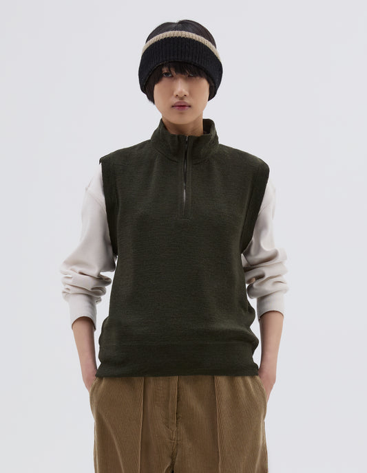 Cropped body image, front view of model wearing dark green wool slipover, with half zip mock turtleneck and flatlock stitch detailing on seams. Styled over cream sweatshirt, with black beanie hat and beige corduroy trousers.