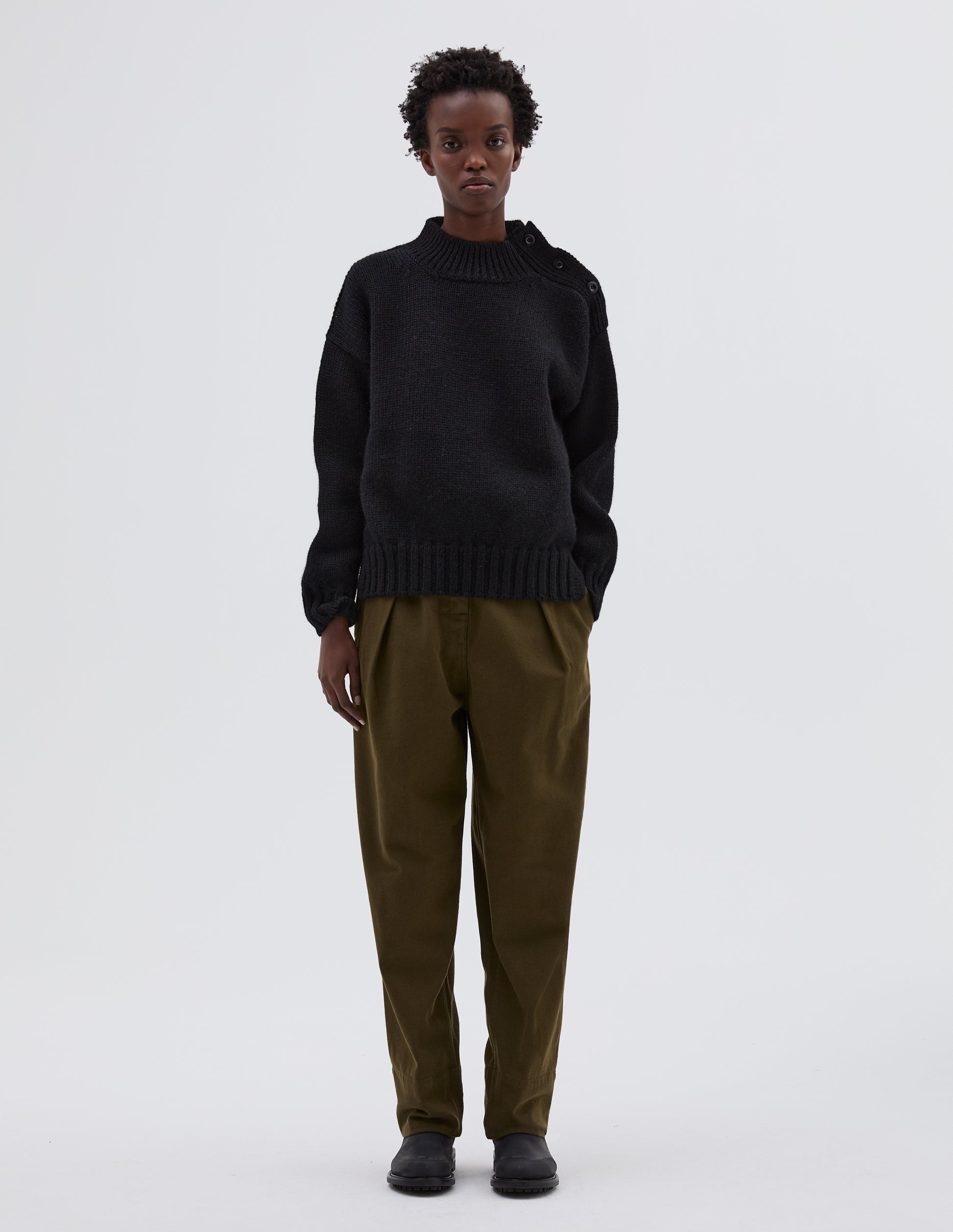 Full body image, front view model wearing black high neck jumper with corozo button detailing on neck and contrast rib on neck, cuffs and hem. Styled with green trousers and black leather boots.