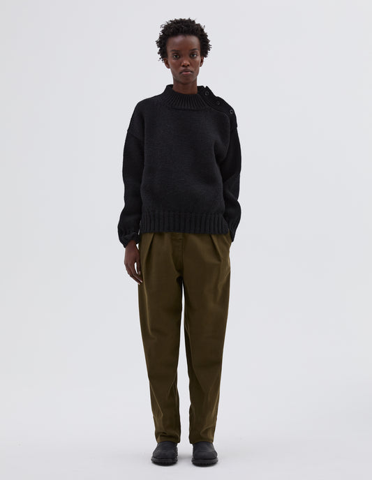 Full body image, front view model wearing black high neck jumper with corozo button detailing on neck and contrast rib on neck, cuffs and hem. Styled with green trousers and black leather boots.