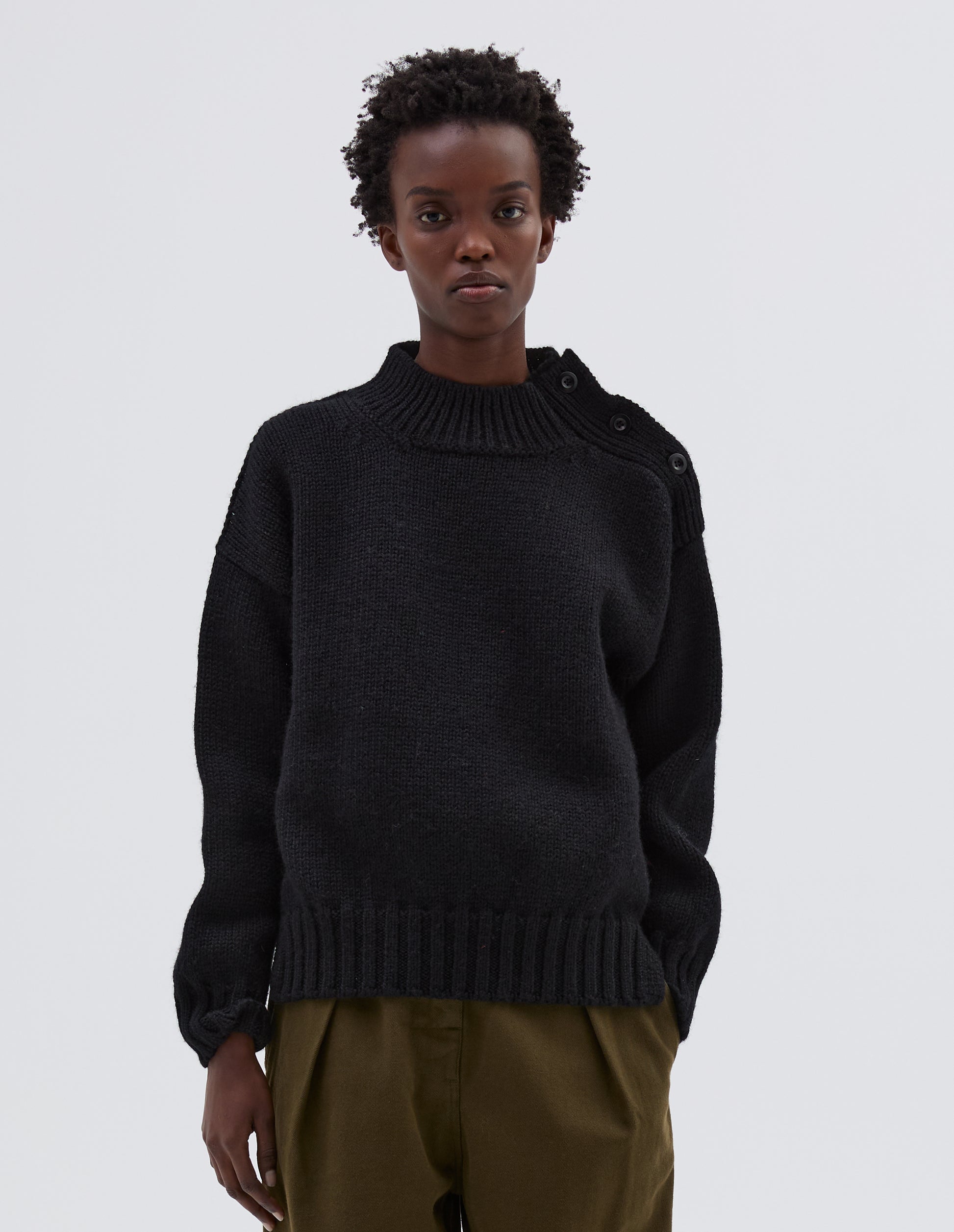 Cropped body image, front view model wearing black high neck jumper with corozo button detailing on neck and contrast rib on neck, cuffs and hem. Styled with green trousers.