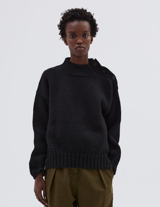 Cropped body image, front view model wearing black high neck jumper with corozo button detailing on neck and contrast rib on neck, cuffs and hem. Styled with green trousers.
