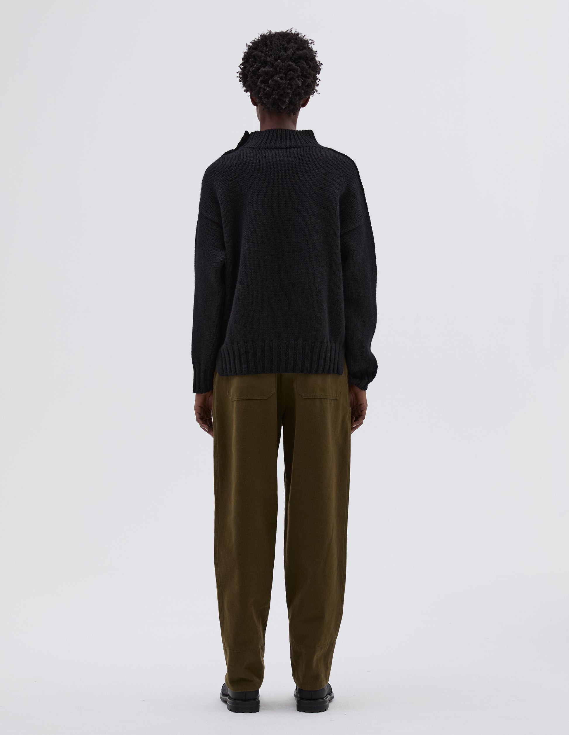 Full body image, back view of model wearing black high neck jumper with contrast rib on neck, cuffs and hem. Styled with green trousers and black leather boots.