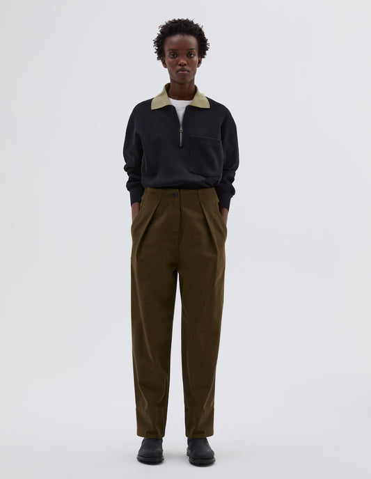 Full body image, front view of model wearing green cotton wool trousers, with zip fly and corozo brace button fastening, self waistband detail, slant pockets, front pleats and deep stitch hem. Styled with black long sleeve polo with contrast collar tucked in, white t shirt and black leather boots.