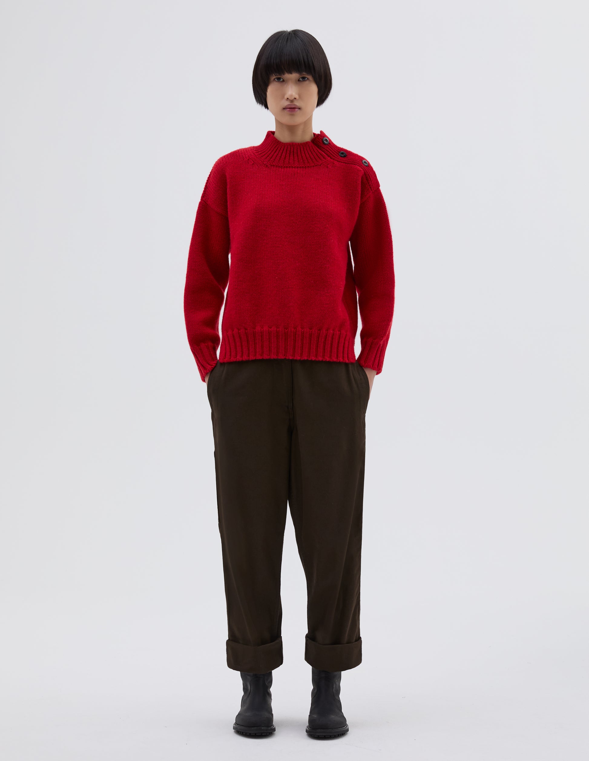 Full body image, front view of model wearing red high neck jumper with corozo button detailing on neck and contrast rib on neck, cuffs and hem. Styled with black trousers and black leather boots.