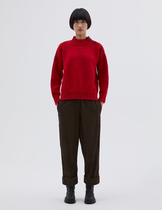 Full body image, front view of model wearing red high neck jumper with corozo button detailing on neck and contrast rib on neck, cuffs and hem. Styled with black trousers and black leather boots.
