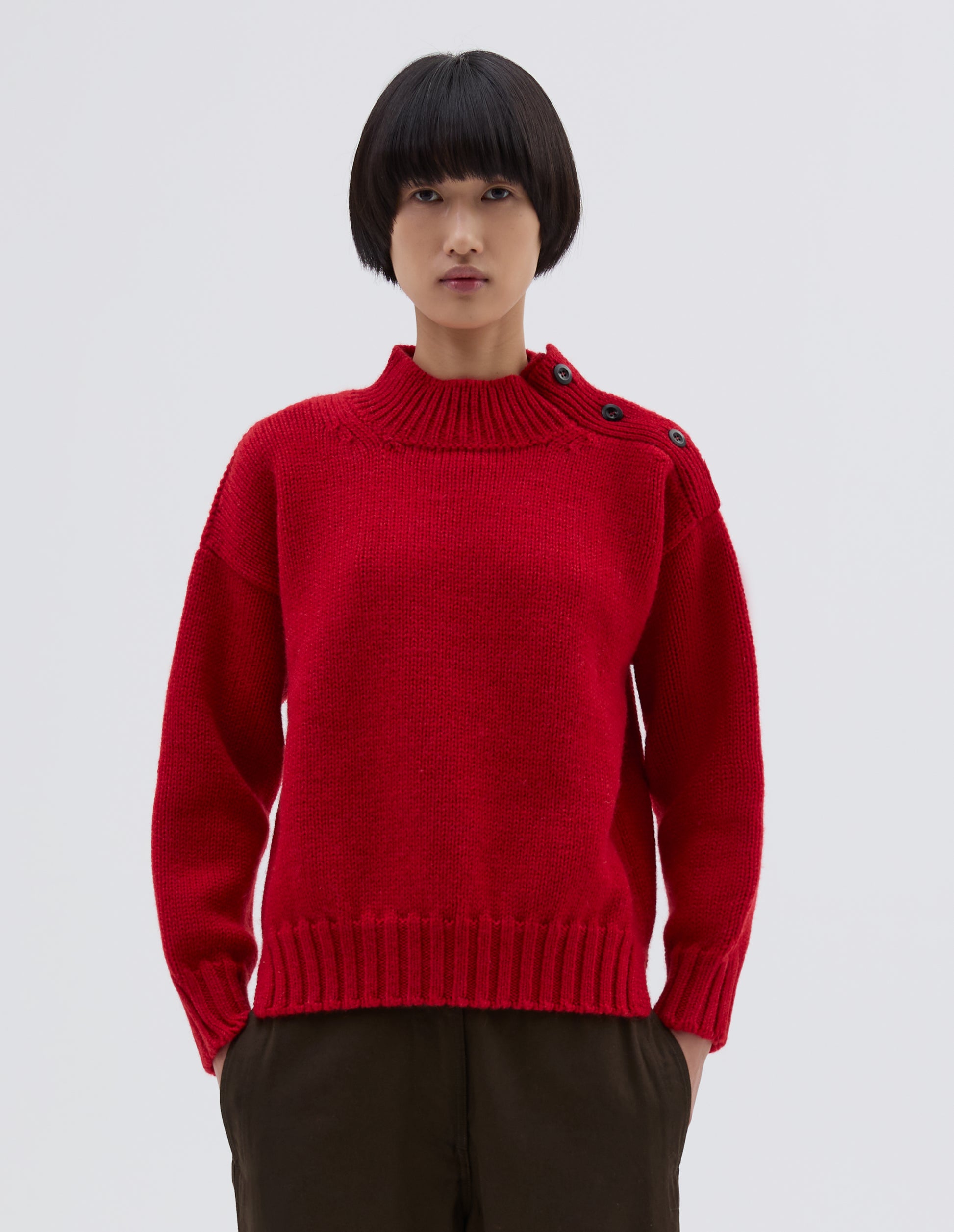 Cropped body image, front view of model wearing red high neck jumper with corozo button detailing on neck and contrast rib on neck, cuffs and hem. Styled with black trousers.