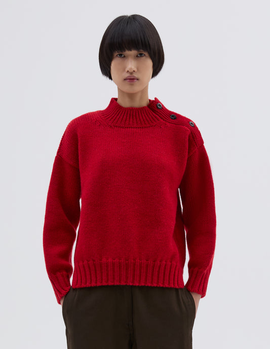 Cropped body image, front view of model wearing red high neck jumper with corozo button detailing on neck and contrast rib on neck, cuffs and hem. Styled with black trousers.