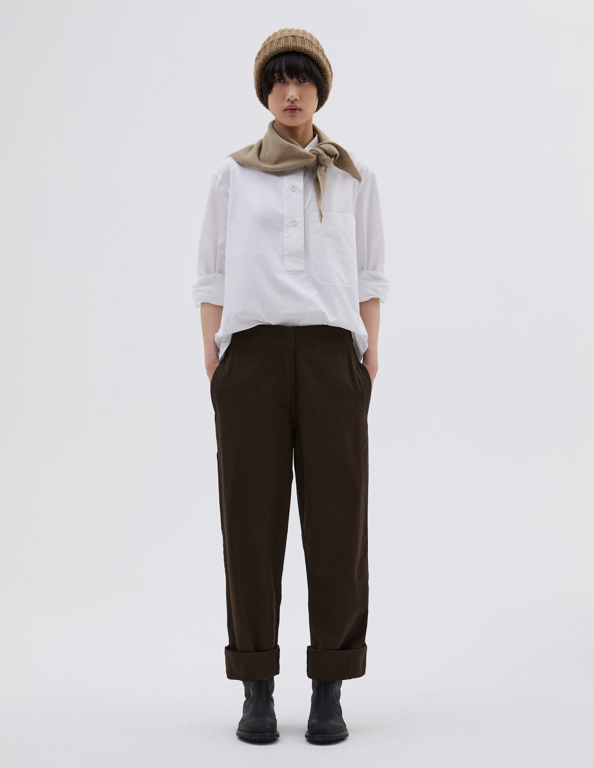 Full body image, front view of model wearing dark brown cotton trousers with zip fly with button through fastening, stitch through waistband, and concealed side pockets. Styled with white shirt tucked in, beige beanie hat, beige knitted scout scarf and black leather boots.