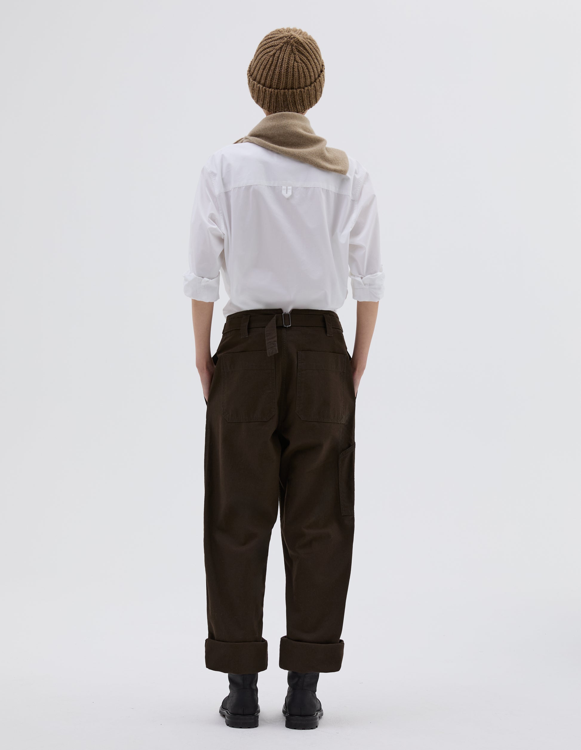 Full body image, back view of model wearing dark brown cotton trousers with stitch through waistband, cinch back fastening with belt loop detail and patch back pockets. Styled with white shirt tucked in, beige beanie hat, beige knitted scout scarf and black leather boots.