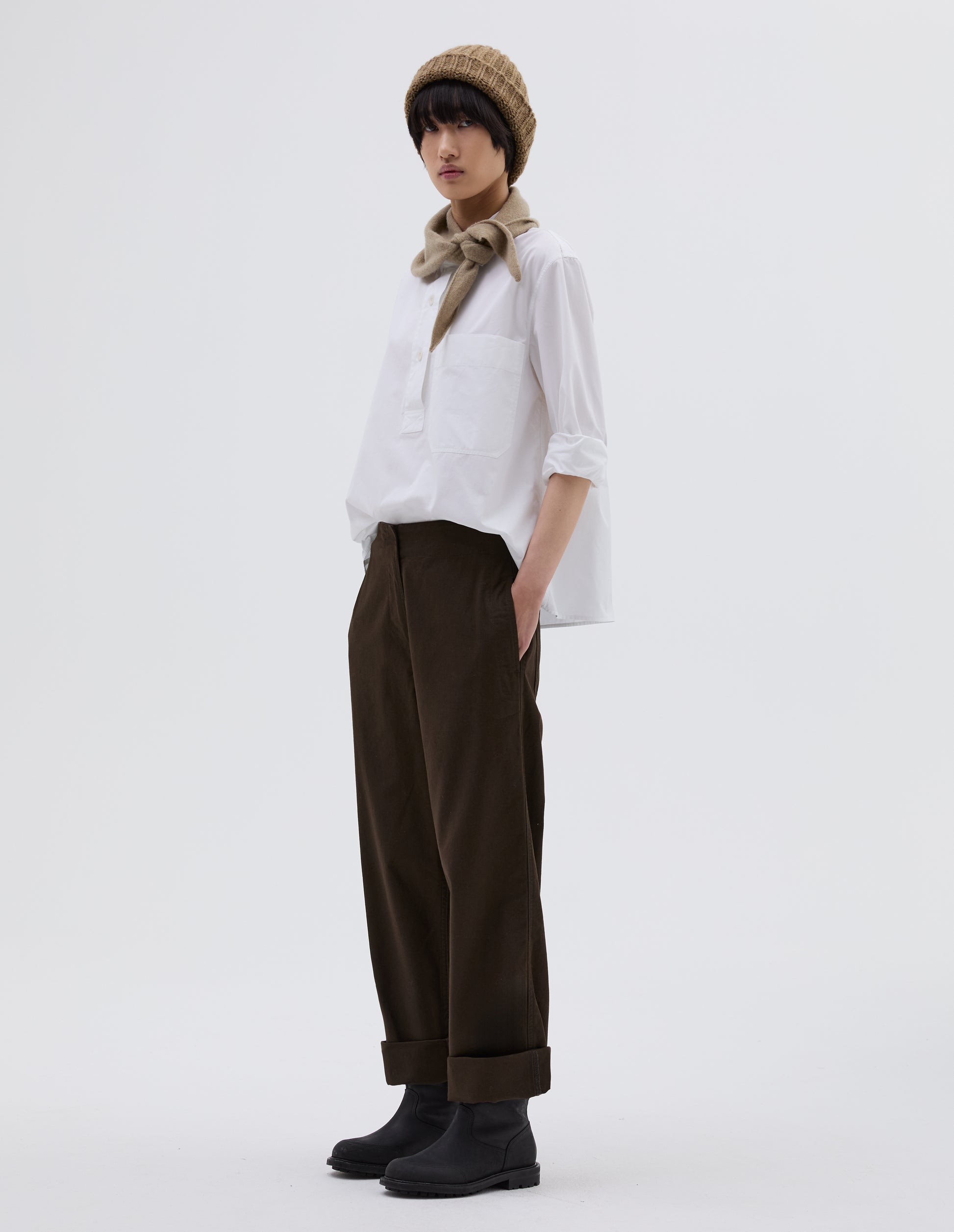 Full body image, side view of model wearing dark brown cotton trousers with zip fly with button through fastening, stitch through waistband, and concealed side pockets. Styled with white shirt tucked in, beige beanie hat, beige knitted scout scarf and black leather boots.