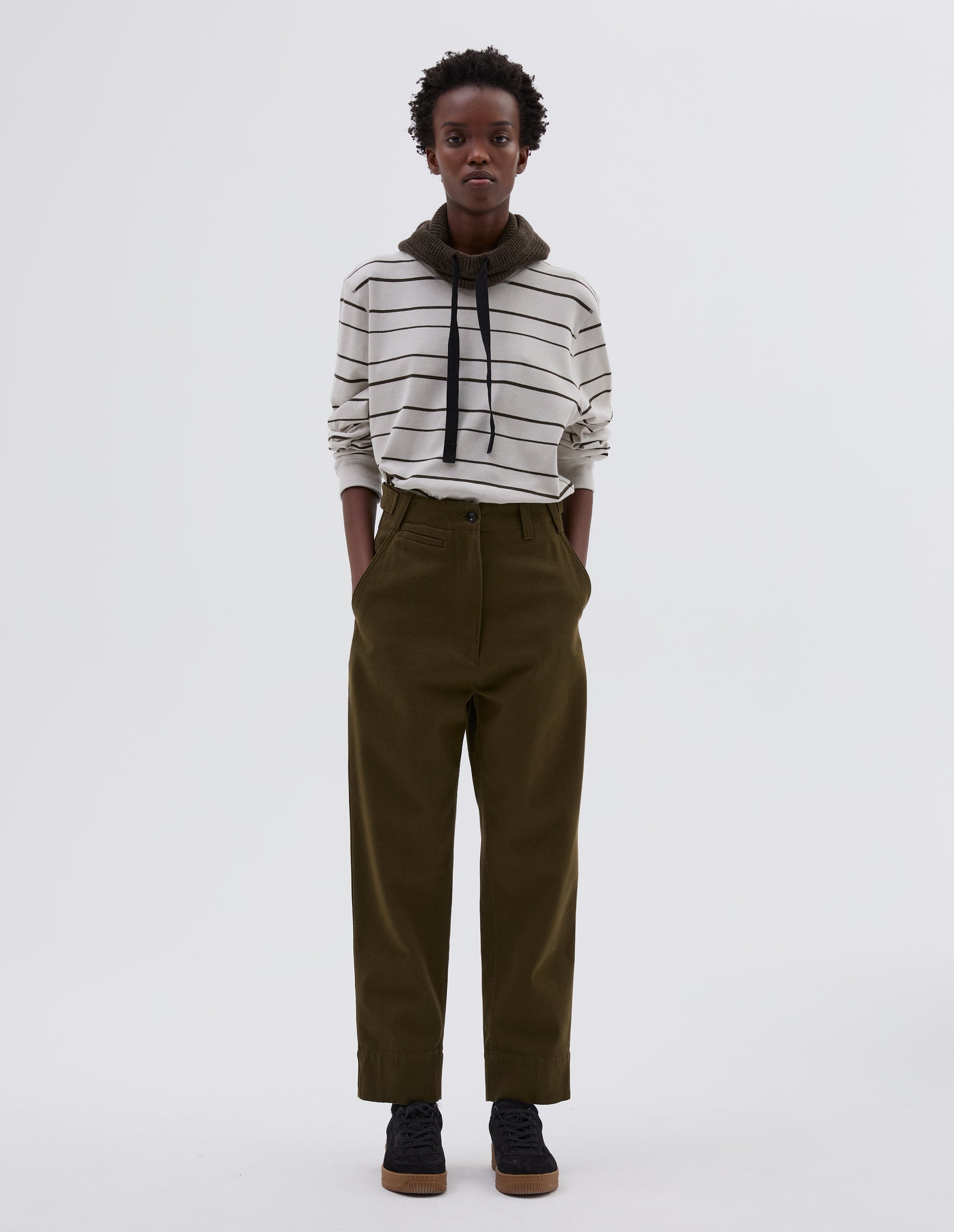 Full body image, front view of model wearing green cotton wool trousers with zip fly button through fastening, button tab side adjusters, belt loops, slant pockets and ticket pocket detail. Styled with white and black striped long sleeve t shirt tucked in, green knitted hood with black herringbone drawcord and black suede trainers.