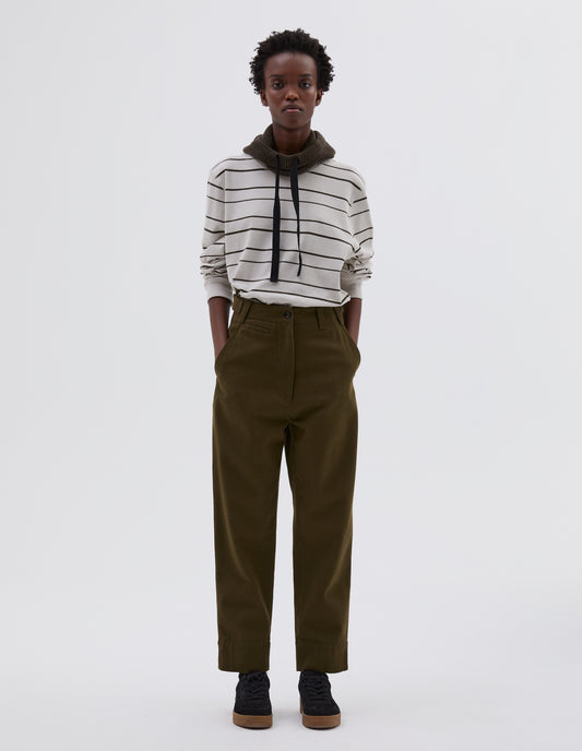 Full body image, front view of model wearing green cotton wool trousers with zip fly button through fastening, button tab side adjusters, belt loops, slant pockets and ticket pocket detail. Styled with white and black striped long sleeve t shirt tucked in, green knitted hood with black herringbone drawcord and black suede trainers.