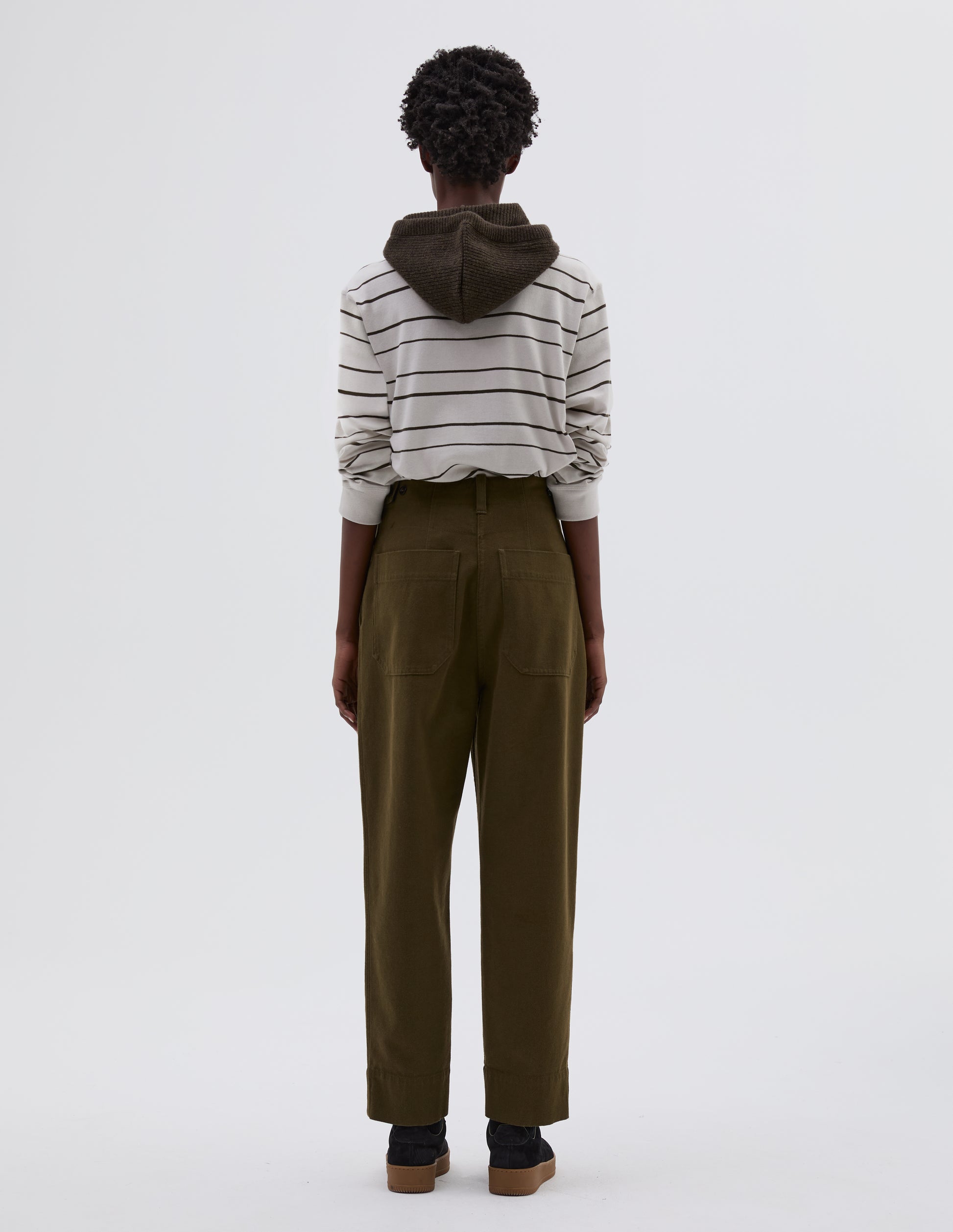 Full body image, back view of model wearing green cotton wool trousers with button tab side adjusters, belt loop and patch back pockets. Styled with white and black striped long sleeve t shirt tucked in, green knitted hood with black herringbone drawcord and black suede trainers.