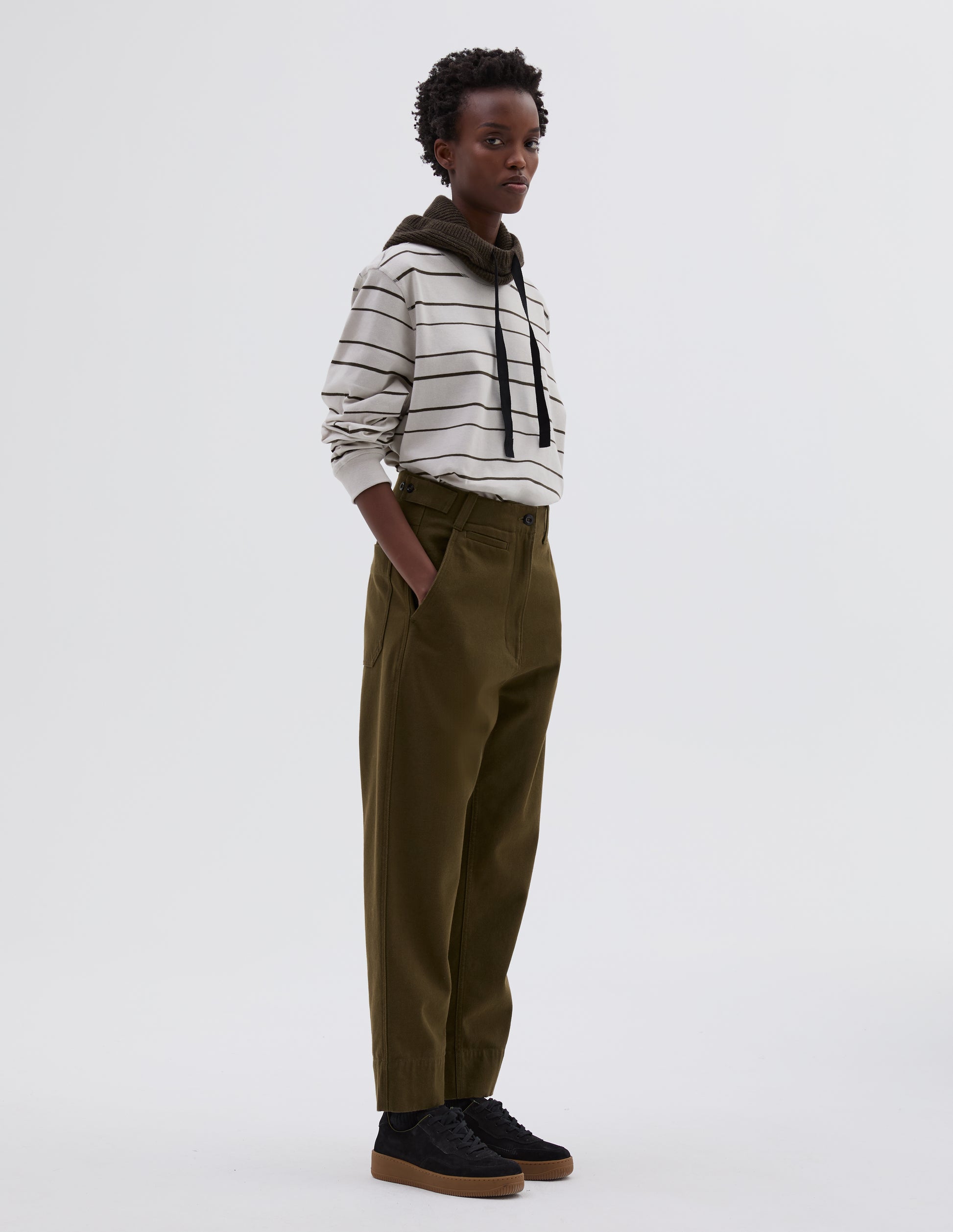 Full body image, side view of model wearing green cotton wool trousers with zip fly button through fastening, button tab side adjusters, belt loops, slant pockets and ticket pocket detail. Styled with white and black striped long sleeve t shirt tucked in, green knitted hood with black herringbone drawcord and black suede trainers.