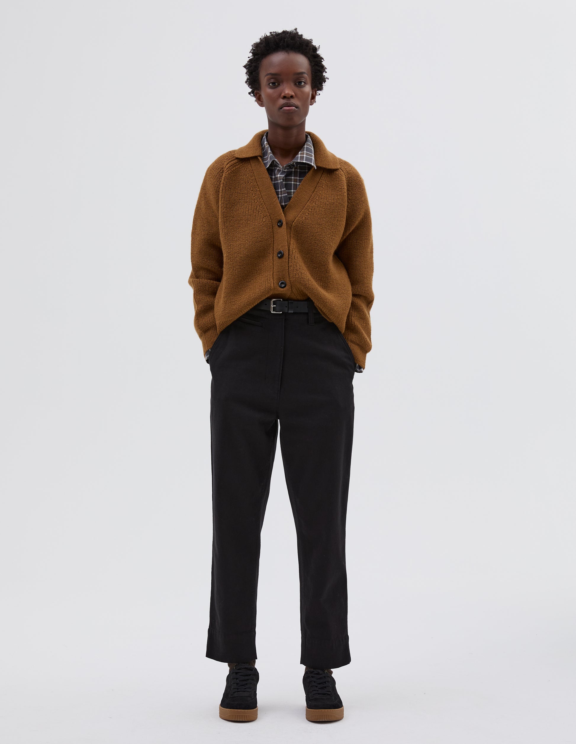 Full body image, front view of model wearing yellow  wool collared cardigan with raglan sleeve, contrast rib on collar, placket and hem and corozo brace button closure. Styled over grey and white checked shirt, brown trousers, black leather belt and black suede shoes.