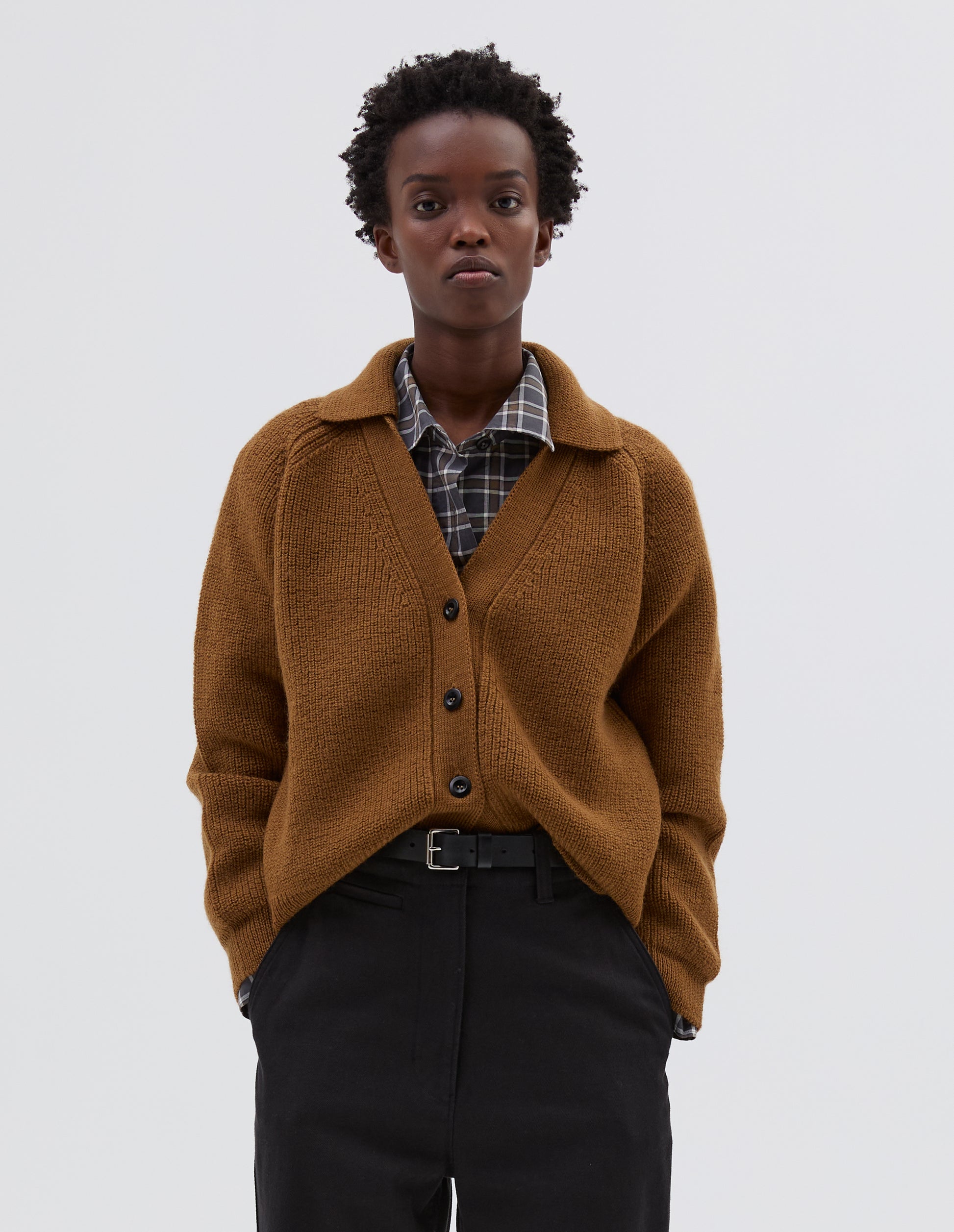 Cropped body image, front view of model wearing yellow  wool collared cardigan with raglan sleeve, contrast rib on collar, placket and hem and corozo brace button closure. Styled over grey and white checked shirt, brown trousers and black leather belt.