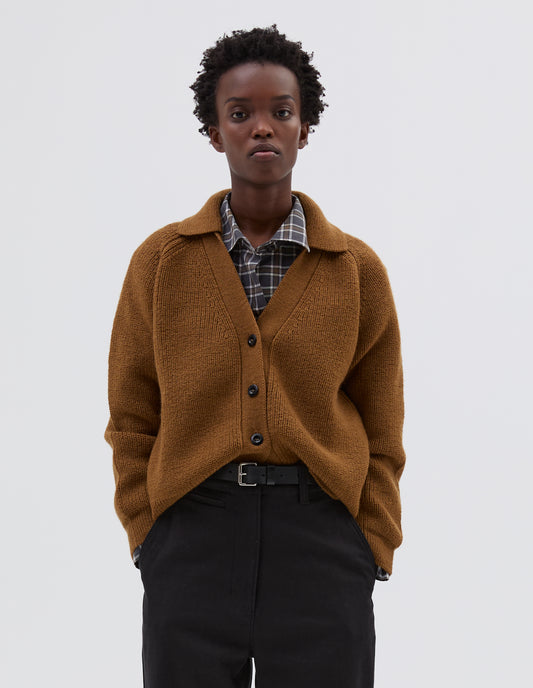 Cropped body image, front view of model wearing yellow  wool collared cardigan with raglan sleeve, contrast rib on collar, placket and hem and corozo brace button closure. Styled over grey and white checked shirt, brown trousers and black leather belt.