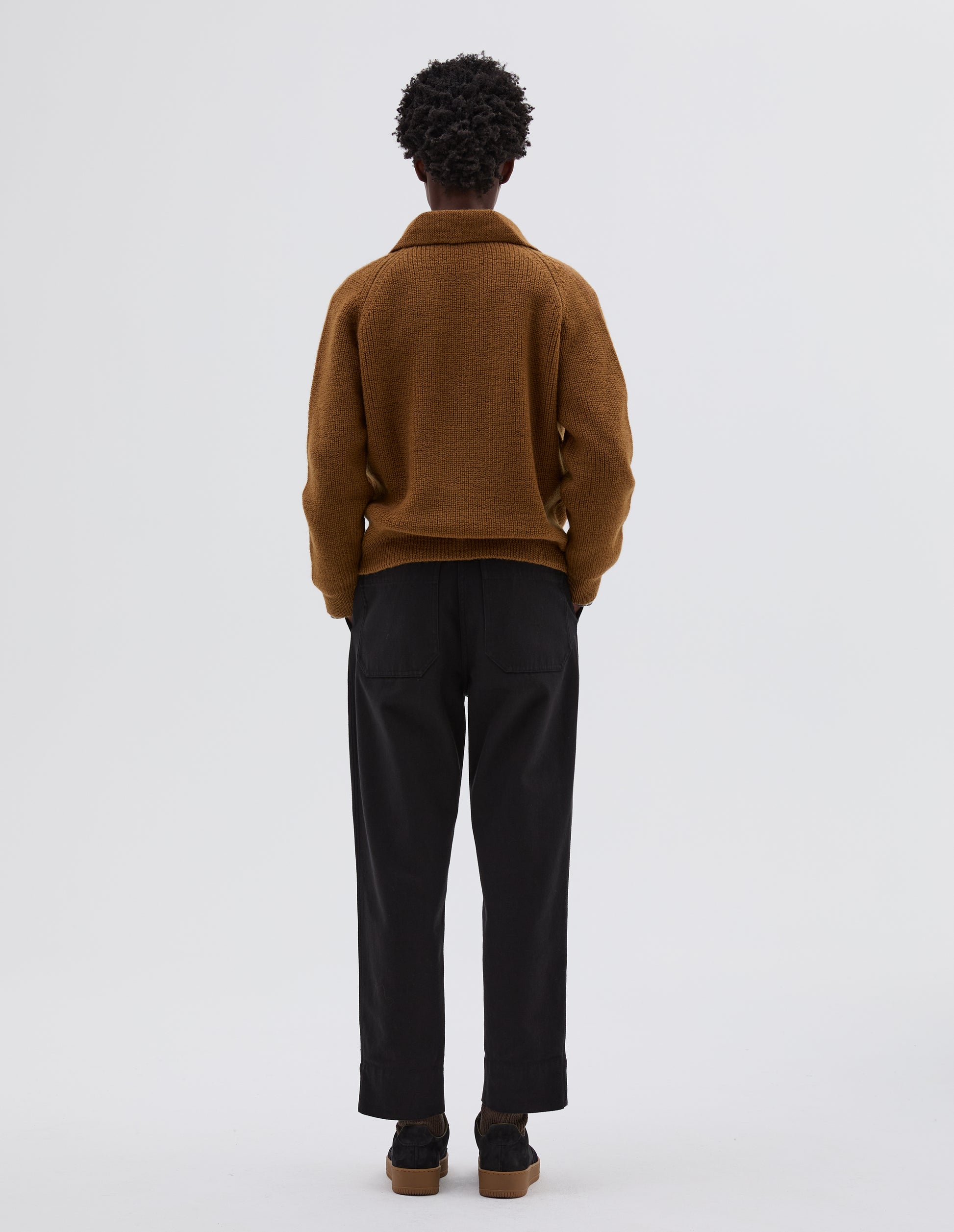 Full body image, back view of model wearing yellow wool collared cardigan with raglan sleeve, contrast rib on collar, placket and hem. Styled with brown trousers, black leather belt and black suede shoes.
