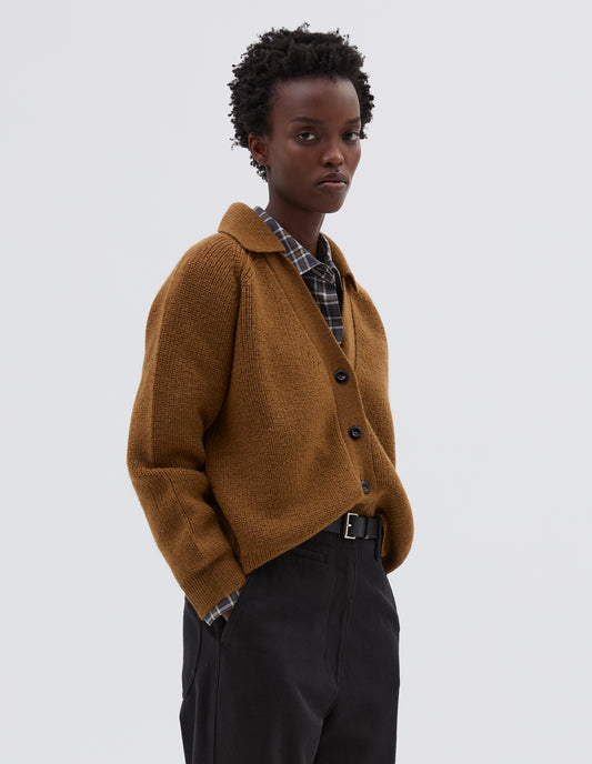 Cropped body image, side view of model wearing yellow  wool collared cardigan with raglan sleeve, contrast rib on collar, placket and hem and corozo brace button closure. Styled over grey and white checked shirt, brown trousers and black leather belt.