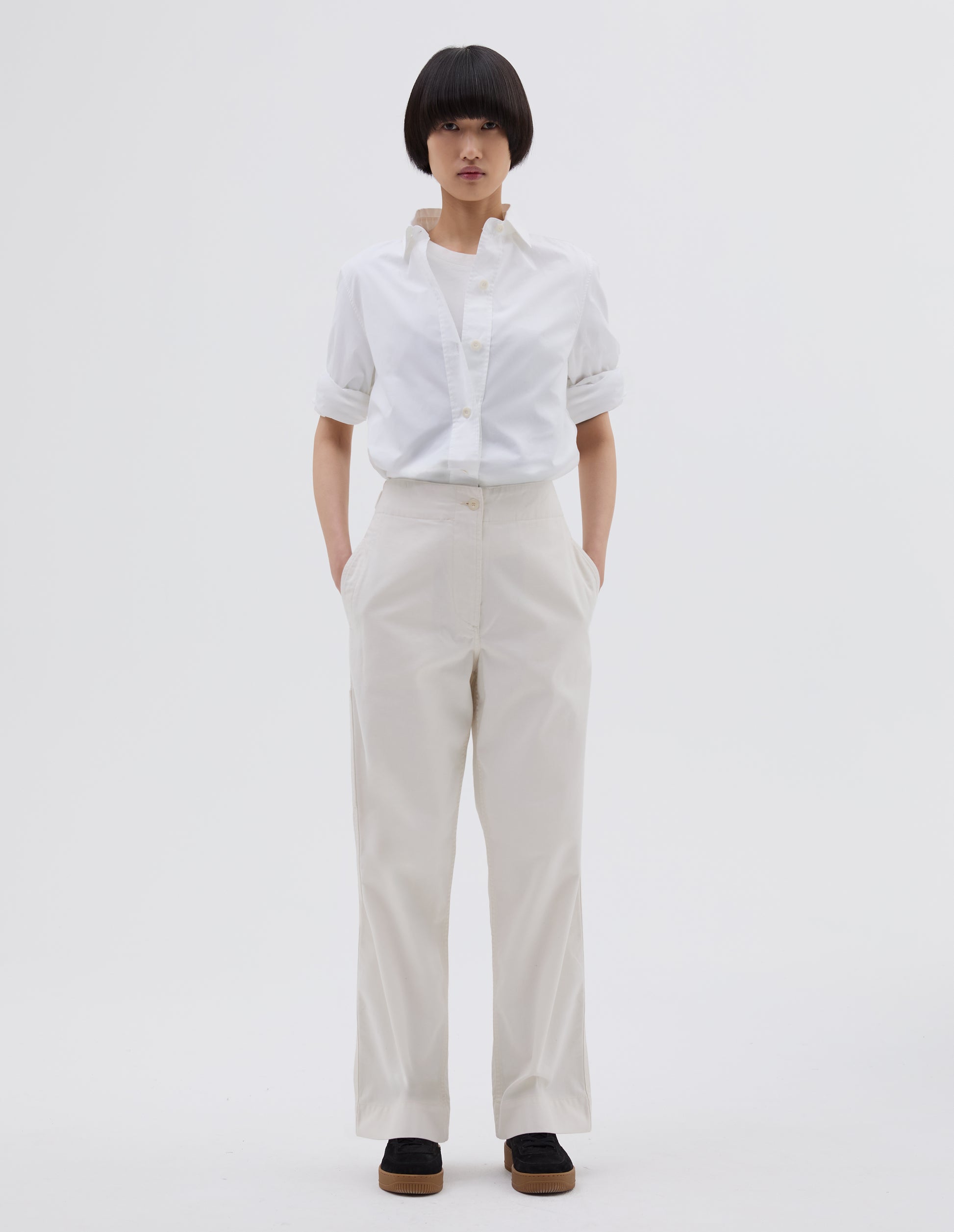 Full body image, front view of model wearing off white cotton trousers with zip fly with button through fastening, stitch through waistband, and concealed side pockets. Styled with white t shirt layered with white shirt tucked in and black suede trainers.