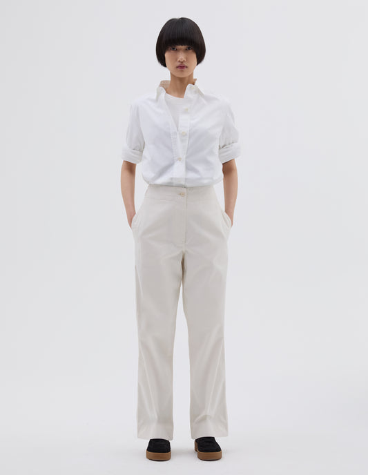 Full body image, front view of model wearing off white cotton trousers with zip fly with button through fastening, stitch through waistband, and concealed side pockets. Styled with white t shirt layered with white shirt tucked in and black suede trainers.