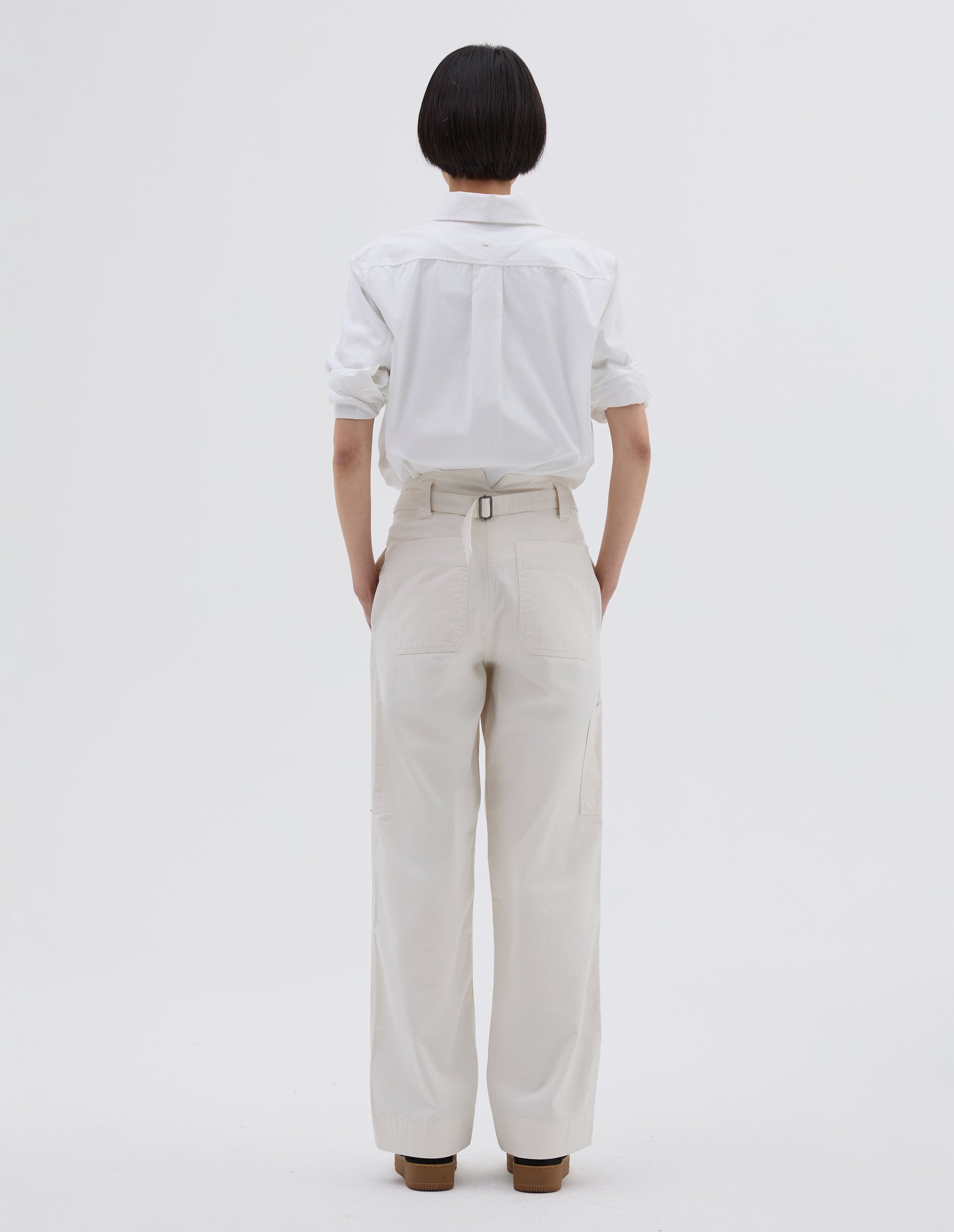 Full body image, back view of model wearing off white cotton trousers with stitch through waistband, cinch back fastening with belt loop detail and patch back pockets. Styled with white shirt tucked in and black suede trainers.