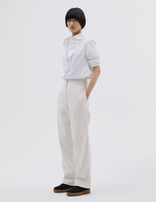 Full body image, side view of model wearing off white cotton trousers with zip fly with button through fastening, stitch through waistband, and concealed side pockets. Styled with white t shirt layered with white shirt tucked in and black suede trainers.