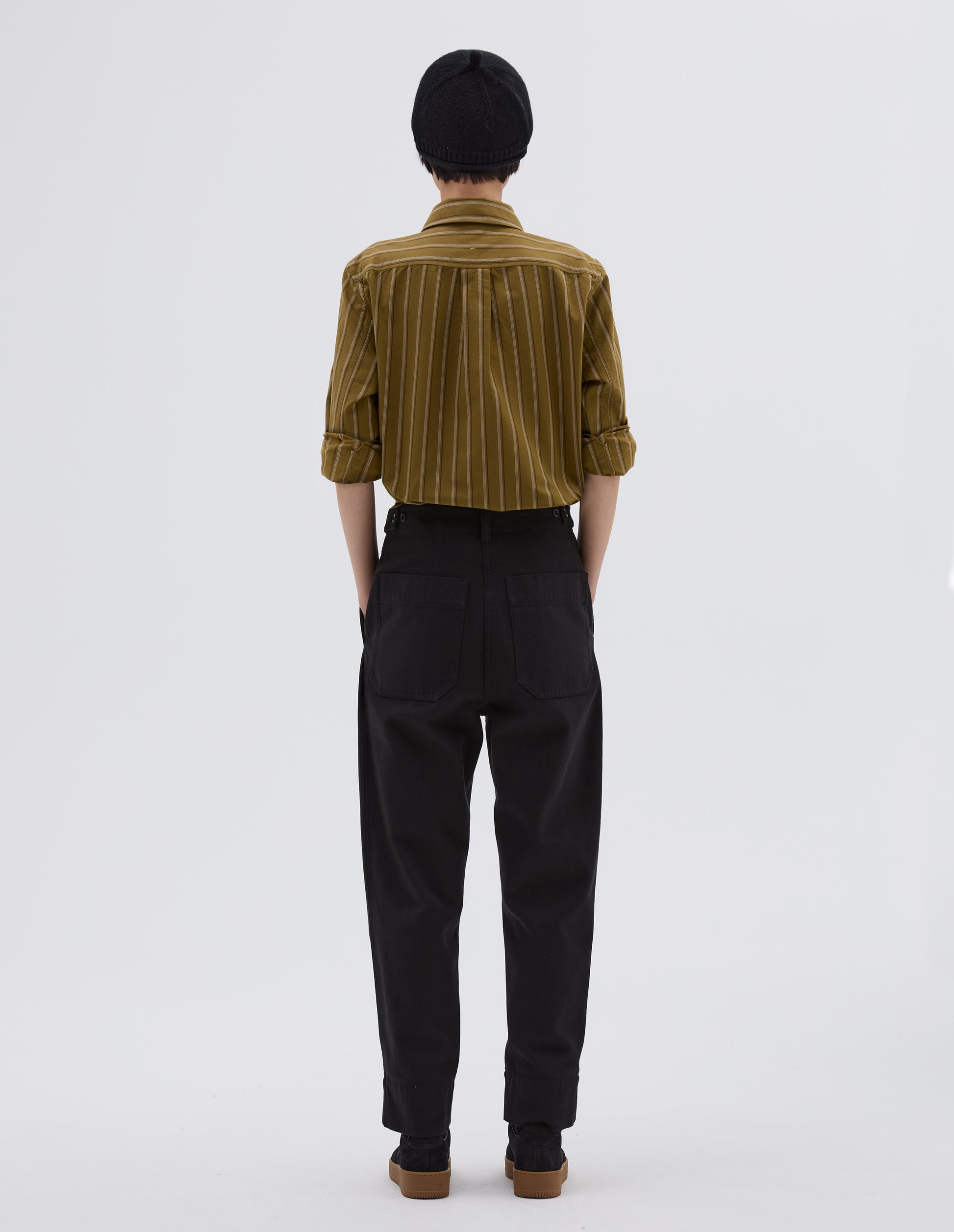 Full body image, back view of model wearing black cotton wool trousers with button tab side adjusters, belt loop and patch back pockets. Styled with yellow, white and black striped shirt tucked in, black beanie hat and black suede trainers.