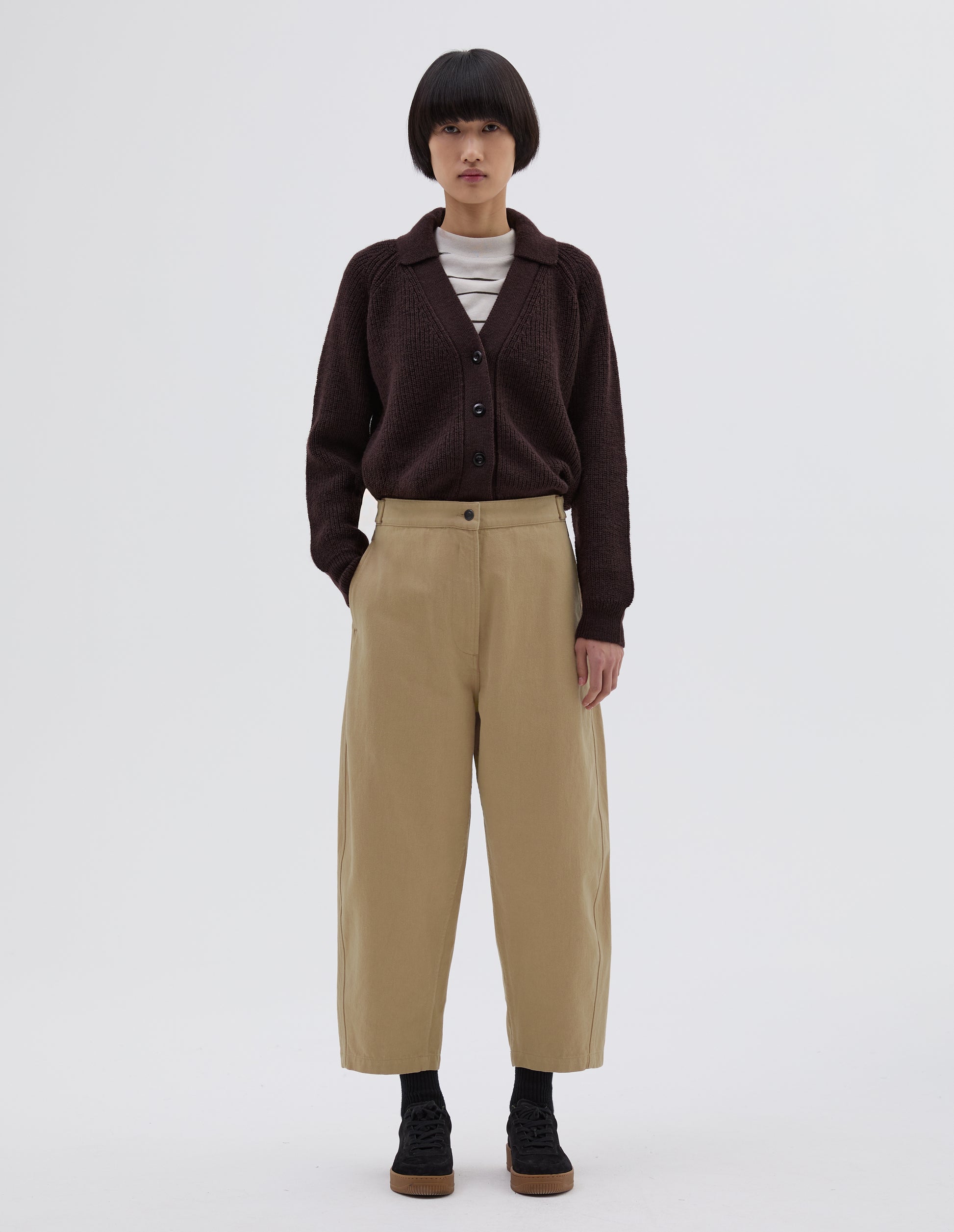 Full body image, front view of model wearing beige trousers with zip fly and button through fastening, side cinch adjusters on waistband and concealed side pockets. Styled with white and black striped long sleeve, dark brown collared cardigan tucked in, black socks and black suede trainers.