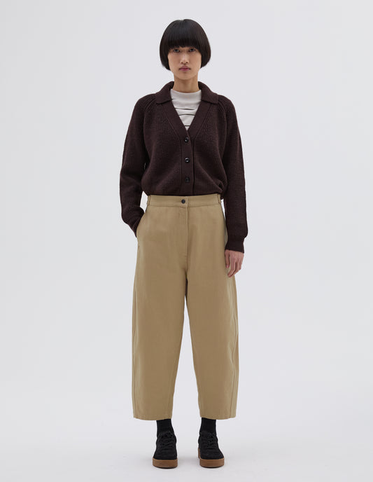Full body image, front view of model wearing beige trousers with zip fly and button through fastening, side cinch adjusters on waistband and concealed side pockets. Styled with white and black striped long sleeve, dark brown collared cardigan tucked in, black socks and black suede trainers.