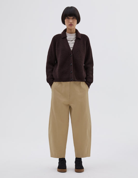 Full body image, front view of model wearing brown  wool collared cardigan with raglan sleeve, contrast rib on collar, placket and hem and corozo brace button closure. Styled over white and black striped long sleeve t shirt, beige trousers and black suede trainers.