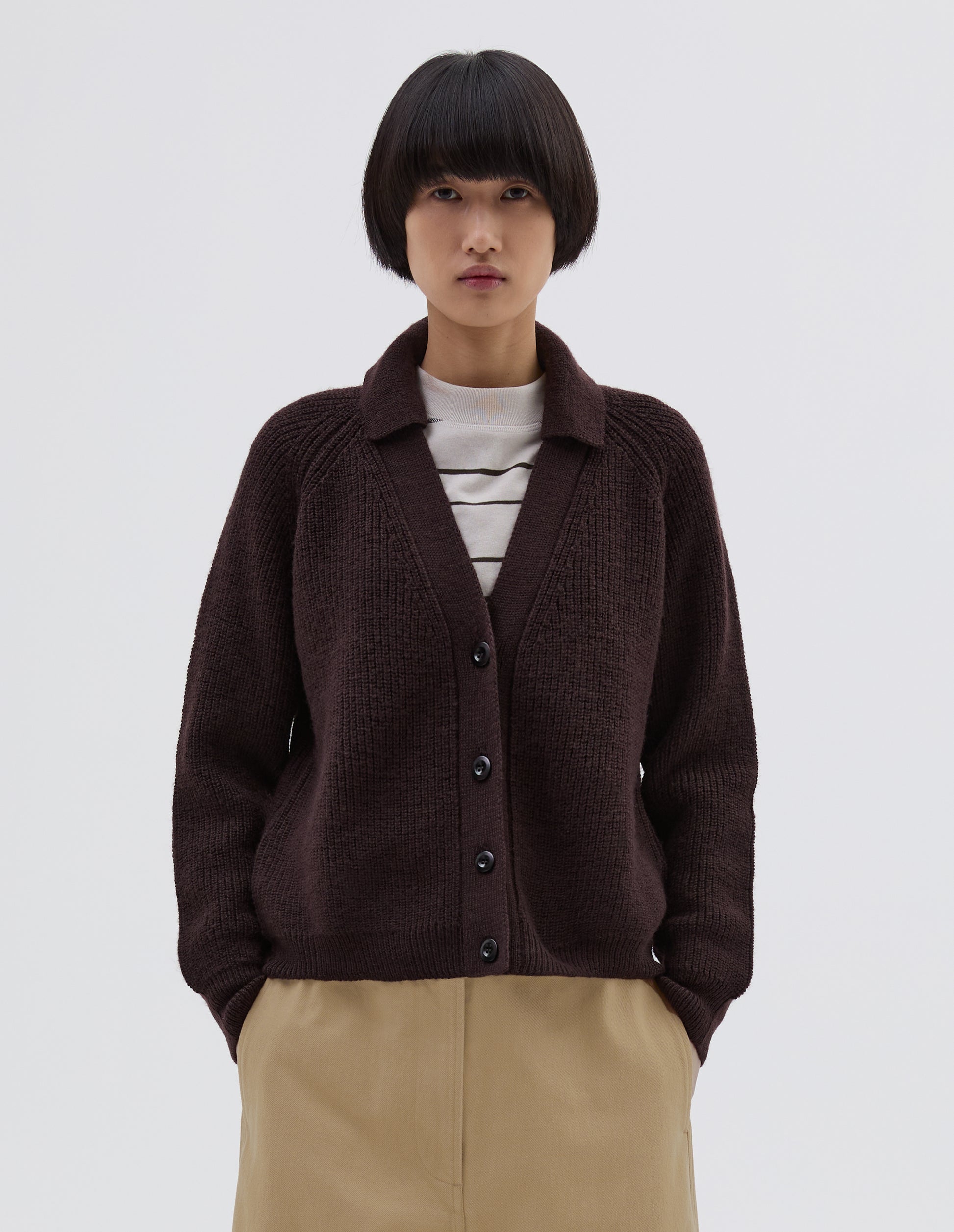 Cropped body image, front view of model wearing brown  wool collared cardigan with raglan sleeve, contrast rib on collar, placket and hem and corozo brace button closure. Styled over white and black striped long sleeve t shirt and beige trousers.