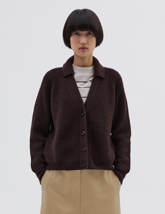 Cropped body image, front view of model wearing brown  wool collared cardigan with raglan sleeve, contrast rib on collar, placket and hem and corozo brace button closure. Styled over white and black striped long sleeve t shirt and beige trousers.