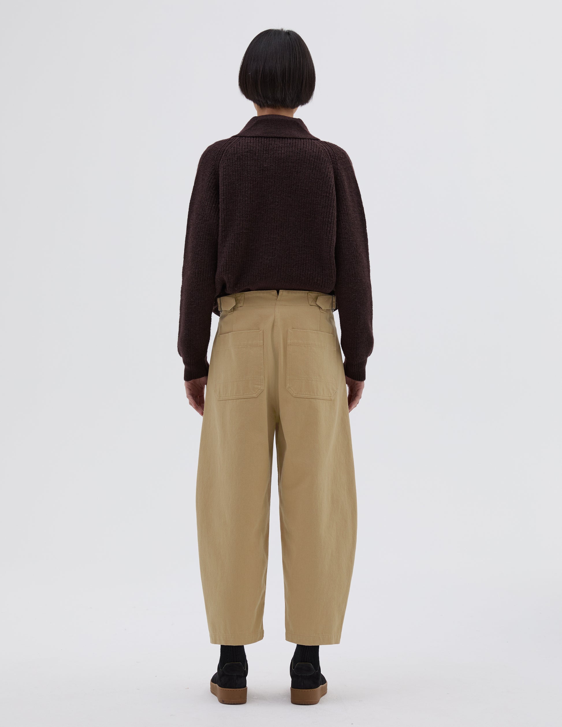 Full body image, back view of model wearing beige trousers with side cinch adjusters on waistband and back patch pockets. Styled with brown collared cardigan tucked in, black socks and black suede trainers.
