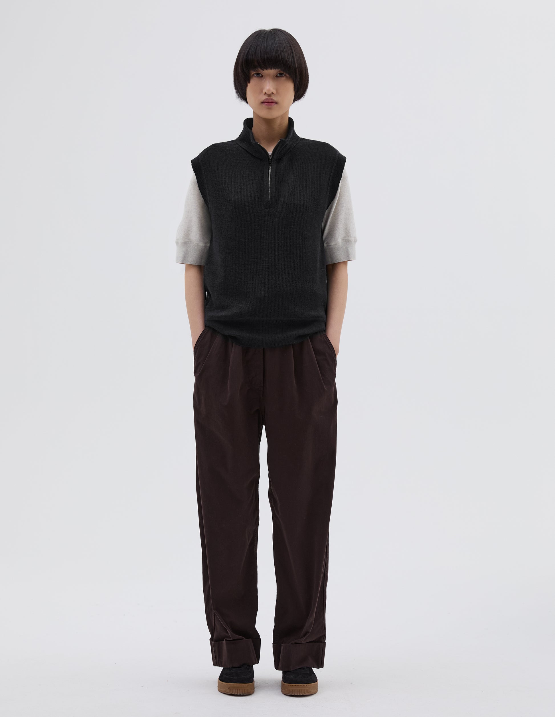 Full body image, front view of model wearing black wool slipover, with half zip mock turtleneck and flatlock stitch detailing on seam. Styled over white t shirt, with brown pleated trousers and black suede trainers.
