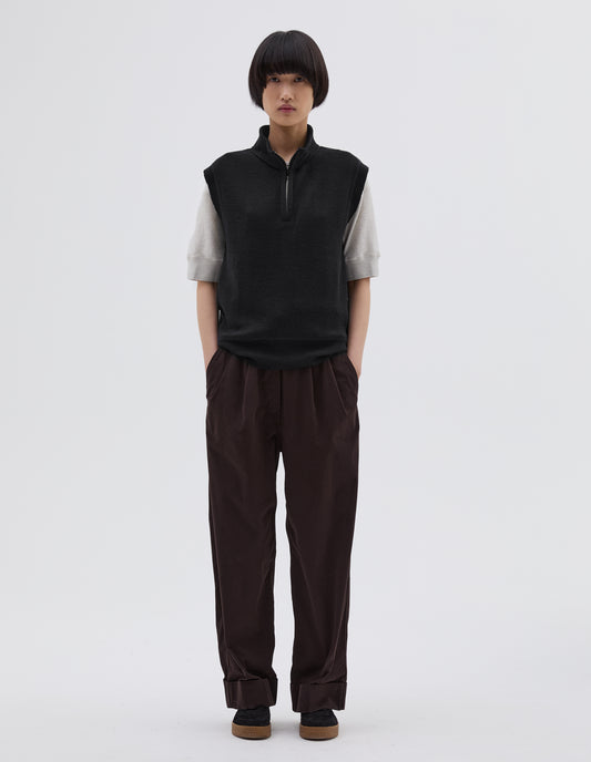 Full body image, front view of model wearing black wool slipover, with half zip mock turtleneck and flatlock stitch detailing on seam. Styled over white t shirt, with brown pleated trousers and black suede trainers.