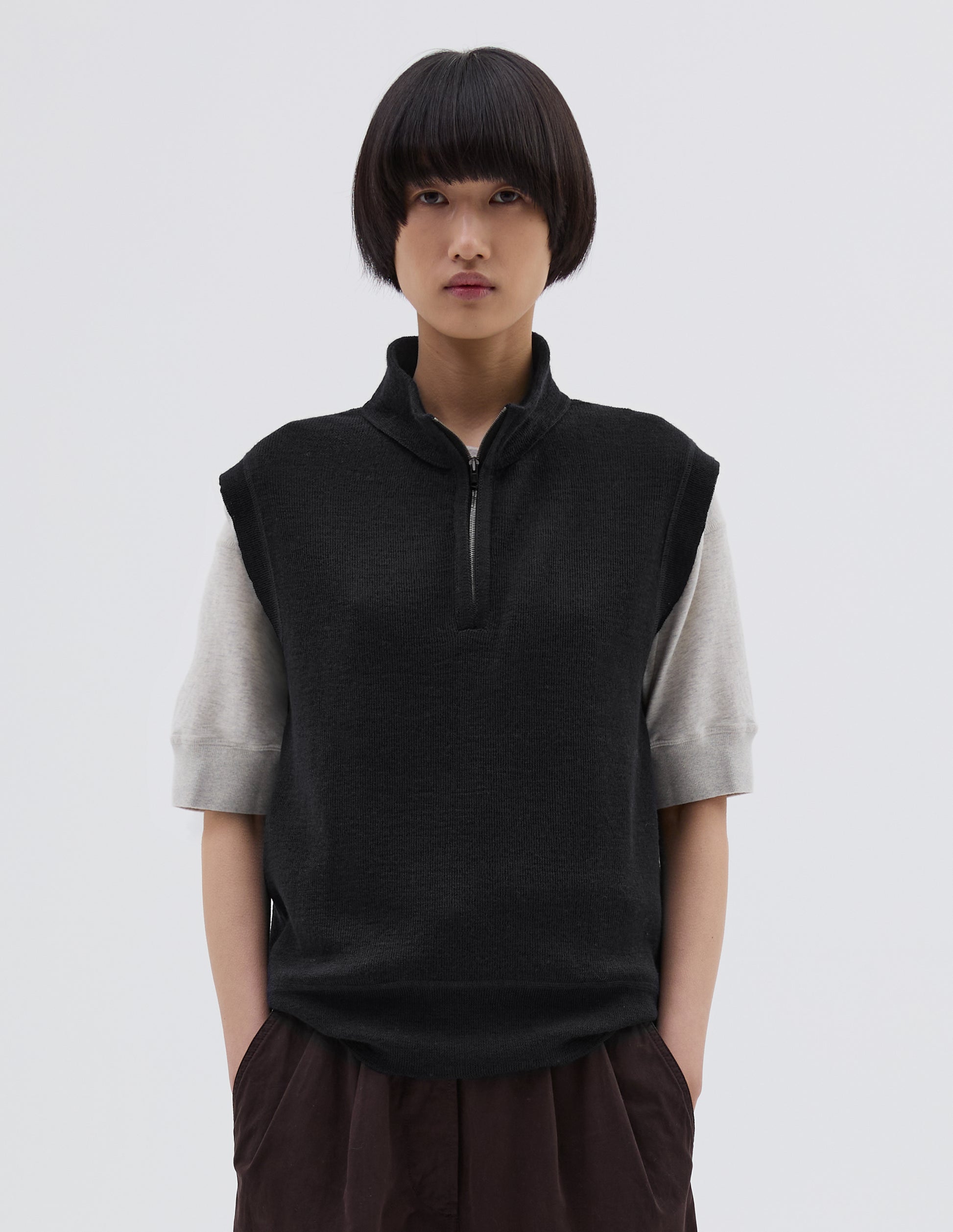 Cropped body image, front view of model wearing black wool slipover, with half zip mock turtleneck and flatlock stitch detailing on seam. Styled over white t shirt, with brown pleated trousers.
