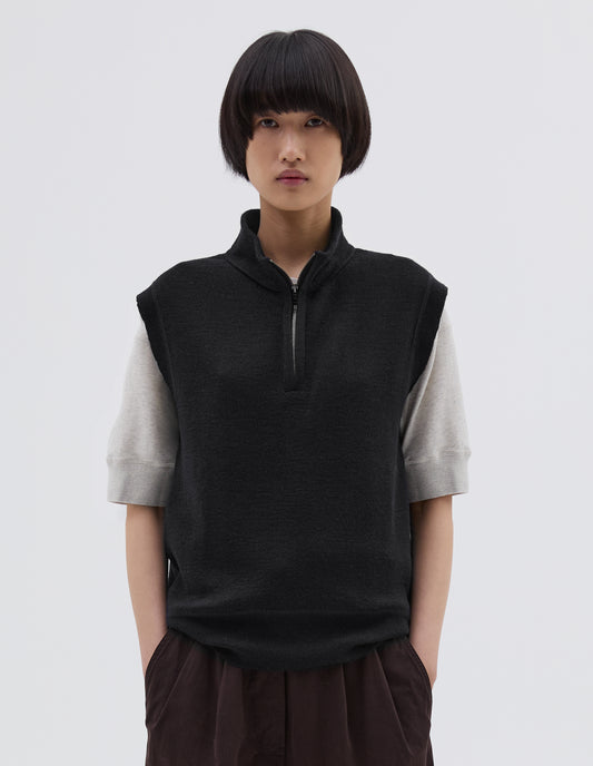 Cropped body image, front view of model wearing black wool slipover, with half zip mock turtleneck and flatlock stitch detailing on seam. Styled over white t shirt, with brown pleated trousers.