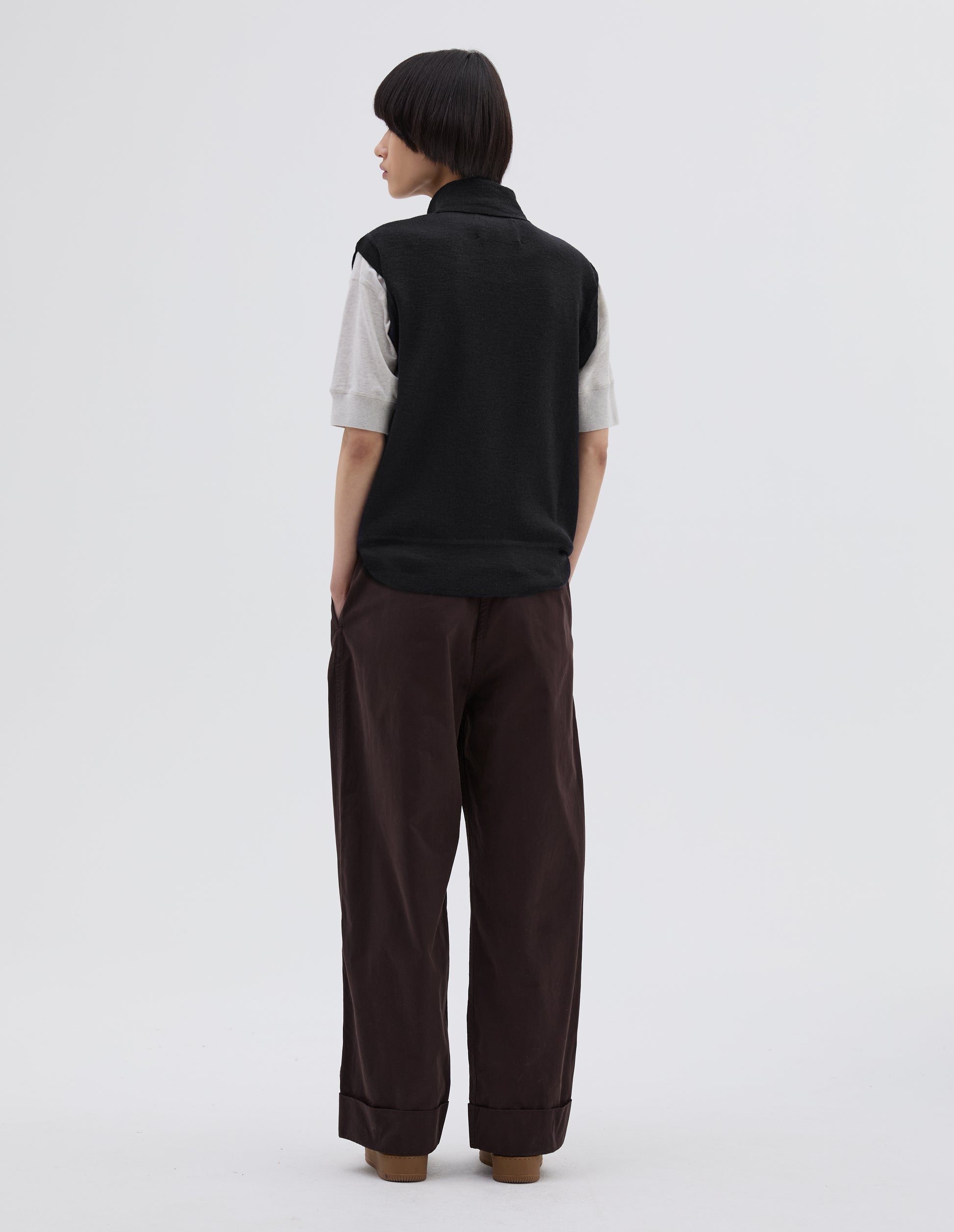 Full body image, back view of model wearing black wool slipover, with half zip mock turtleneck and flatlock stitch detailing on seam. Styled over white t shirt, with brown pleated trousers and black suede trainers.