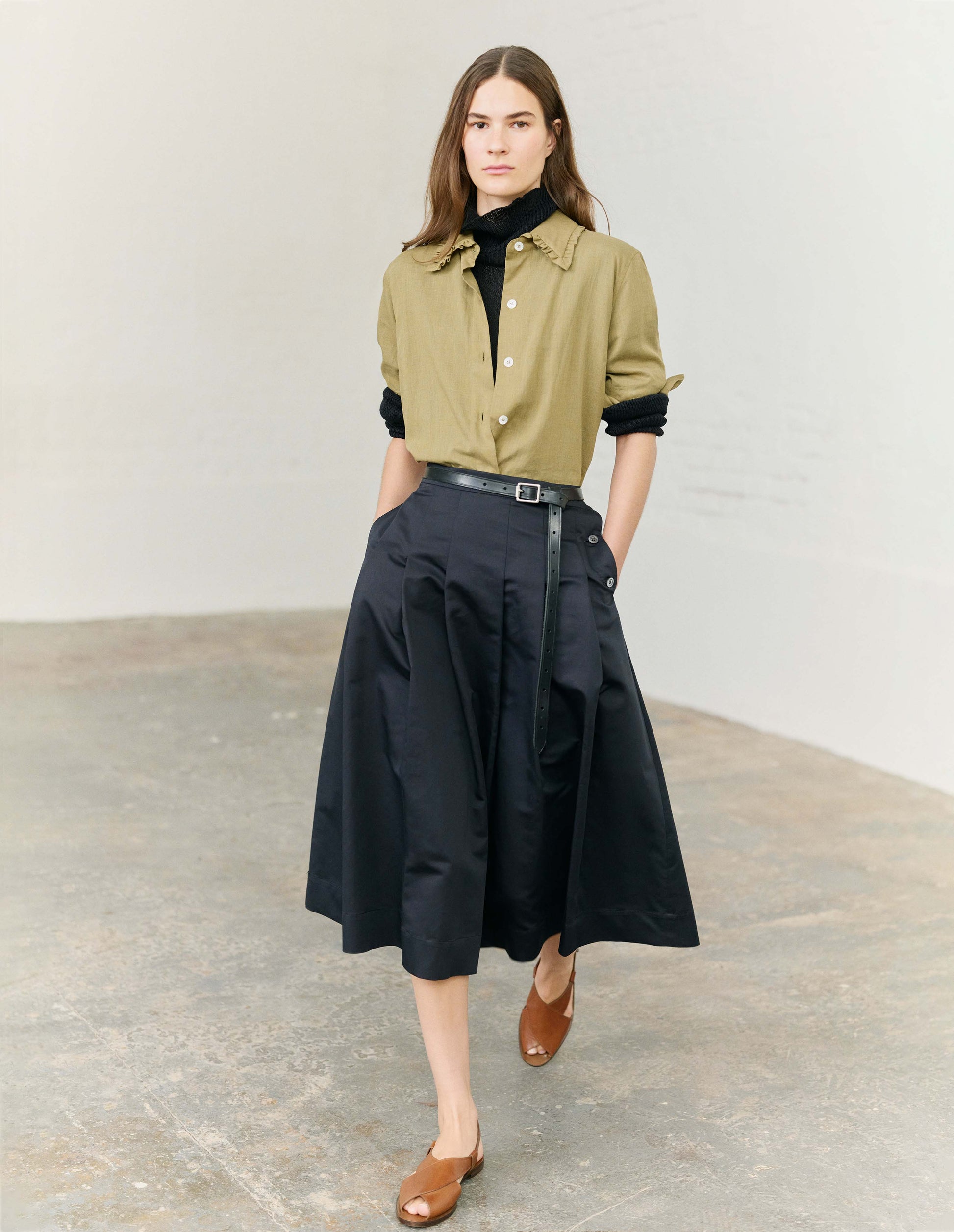 Full body image, front view of model wearing green cotton cashmere frilled collar long sleeve shirt, with horn button closure. Styled with black roll neck jumper, black pleated skirt and light brown leather sandals.