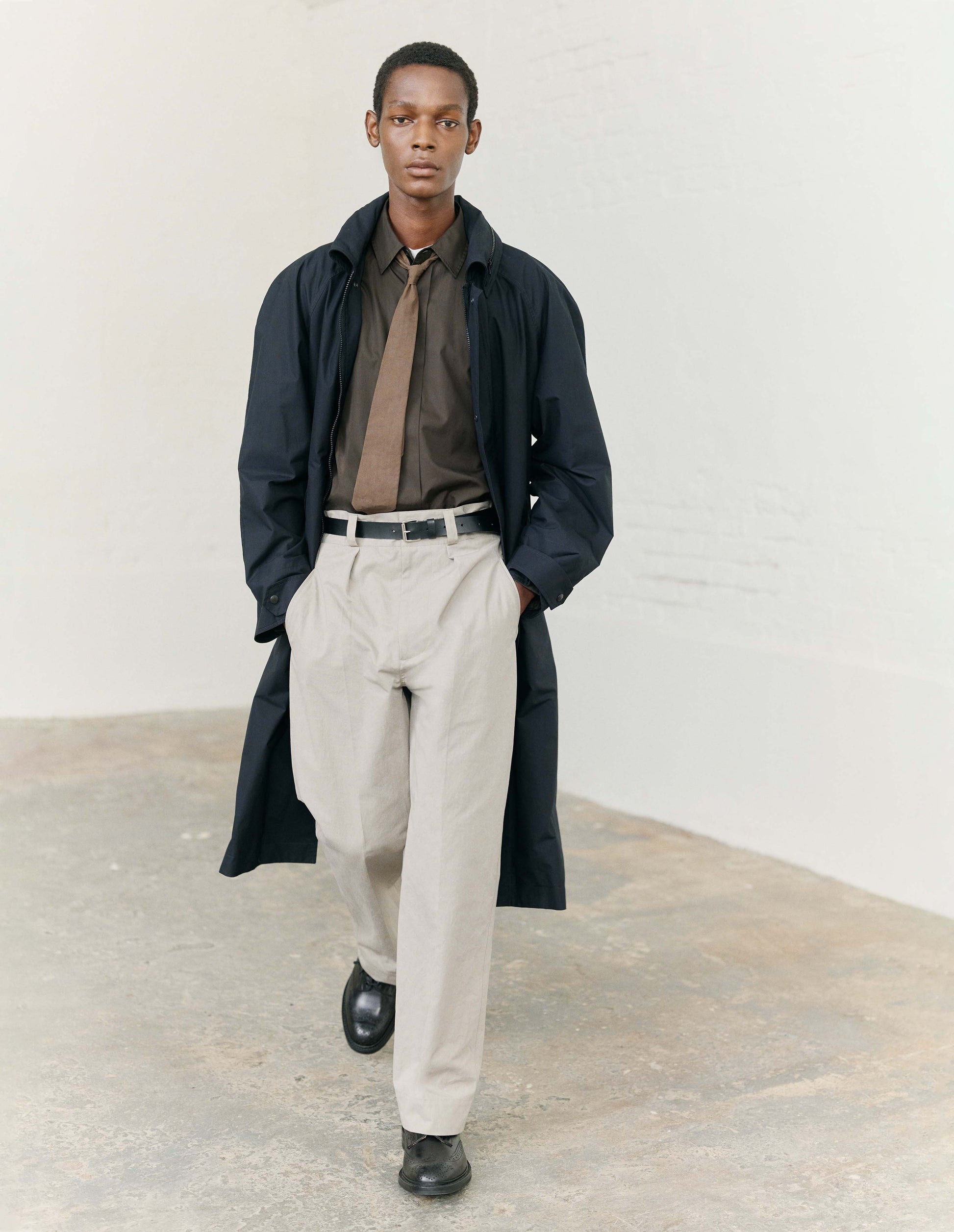 Full body image, front view of model wearing light grey cotton trousers, with single pleats, horn button closure, slant jet pockets and belt loops. Styled with green shirt tucked into waistband, with light brown tie, dark navy blue coat and black leather shoes. 