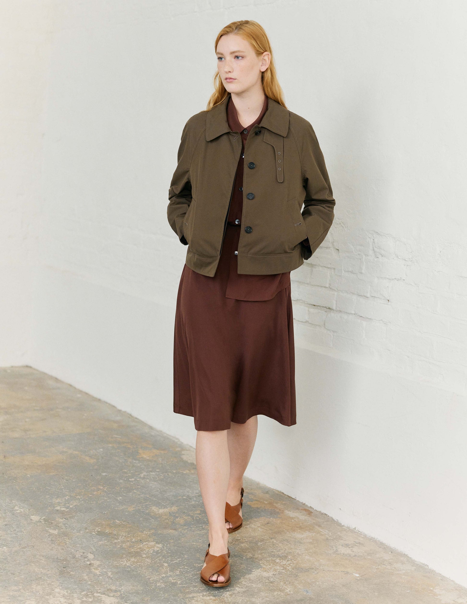Full body image, front view of model wearing greenish brown ventile cotton poplin long sleeve short trench, with horn button closure, slant side pockets, with removable storm tab and hanger loop on collar. Styled with brown collared shirt and matching brown skirt and light brown leather sandals.