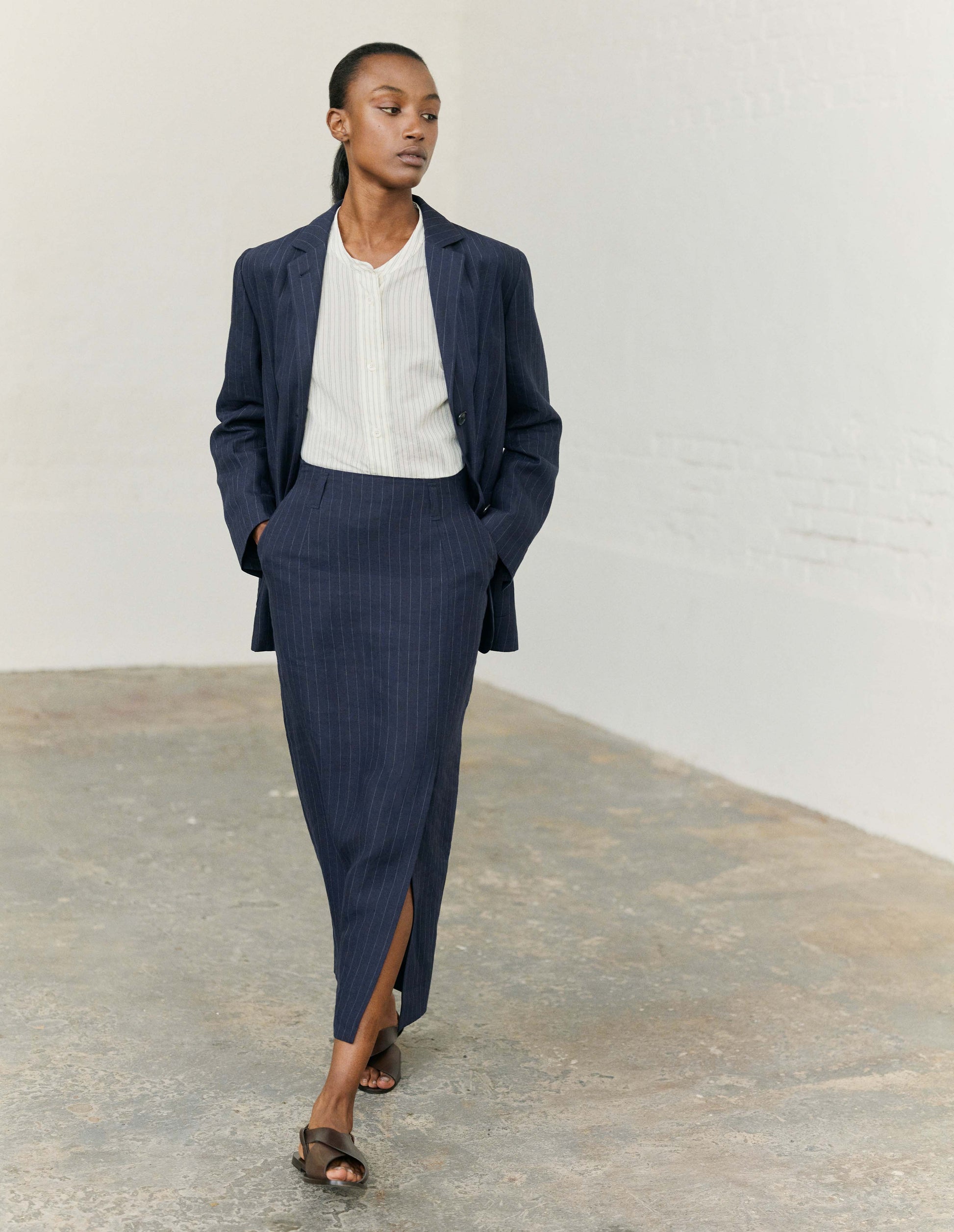 Full body image, front view of modelw wearing navy and grey pinstripe linen skirt, with offset front vent, concealed side zip fastening, slant side pockets and belt loops. Styled with matching navy and grey pinstripe blazer and collarless white and grey pinstripe shirt with dark brown leather sandals.