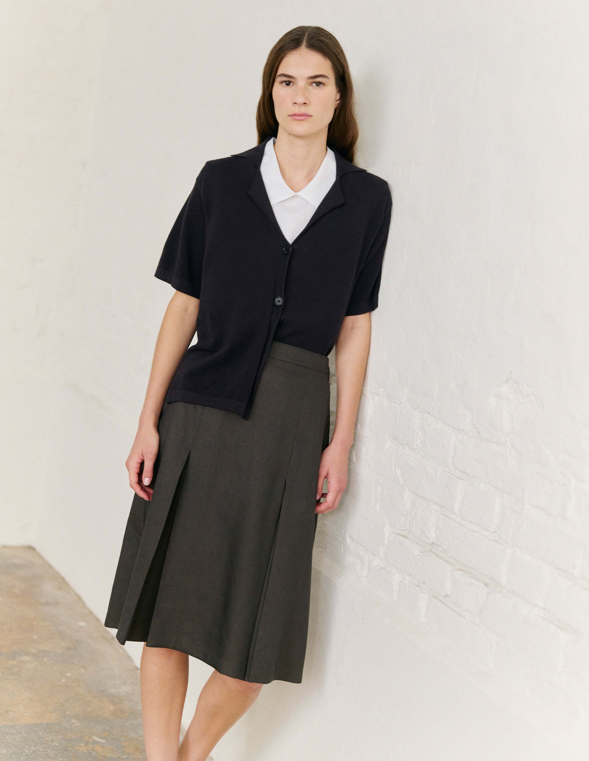 Full body image, front view of model wearing greenish brown regular A line knee length skirt, minimal waistband with full hem. Styled with white short sleeve shirt and black short sleeve knitted shirt.