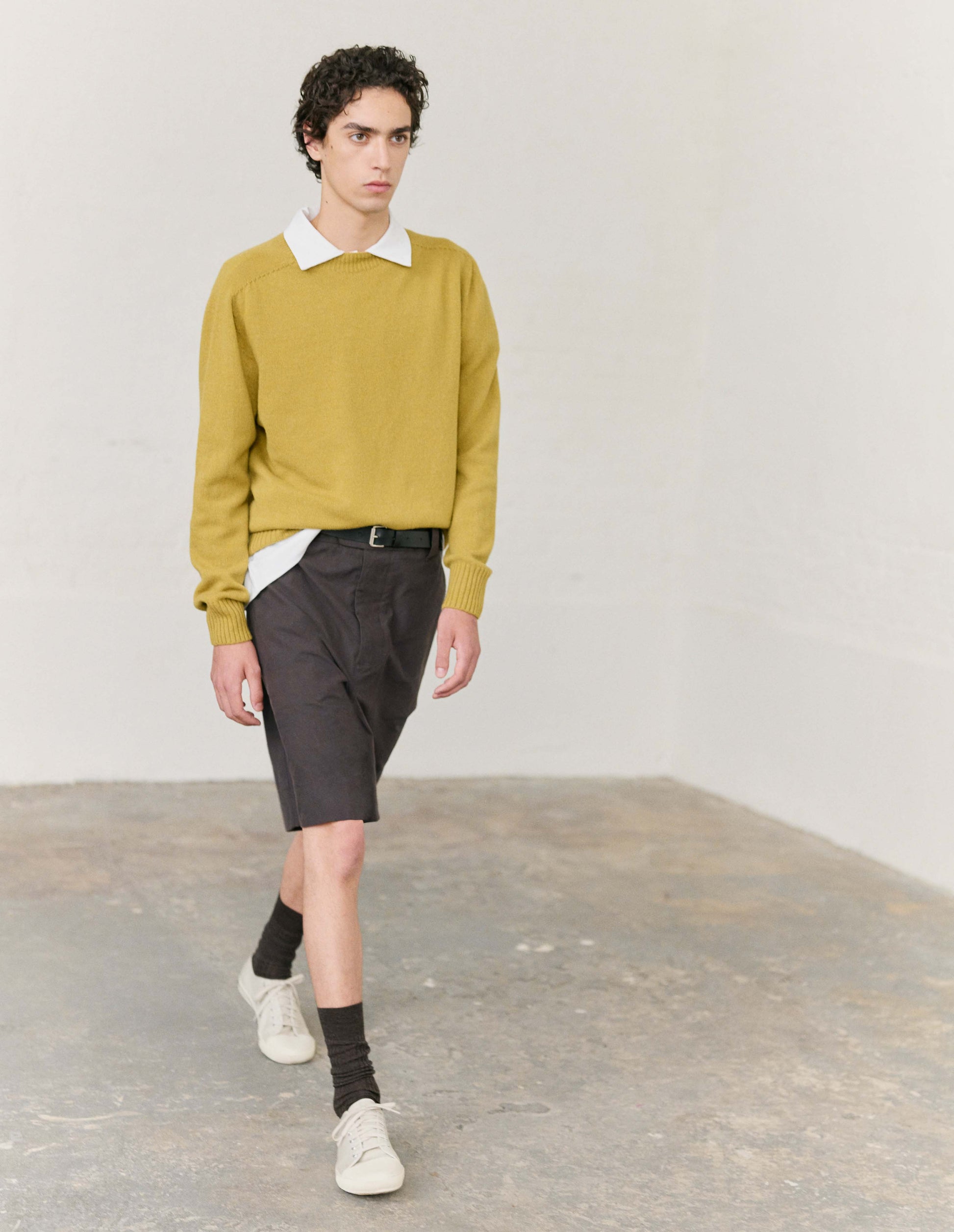 Full body image, front view of model wearing dark brown shorts with button fly and button tap fastening. Styled with white collared shirt and yellow jumper tucked in, with brown socks and white canvas trainers. 