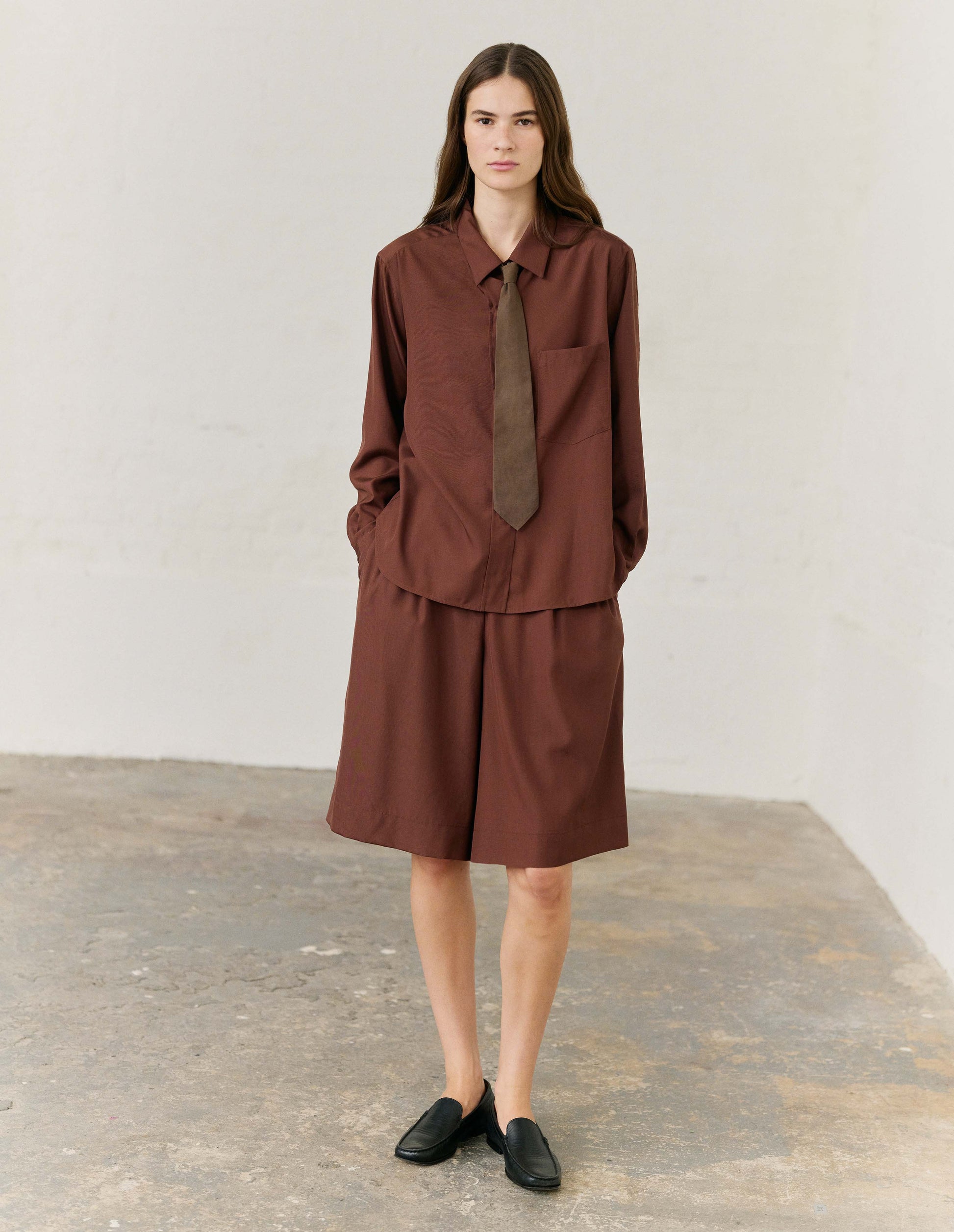 Full body image, front viBrown long shorts with double pleats, hook and bar tab fastening waistband, zip fly and slant side pocketsew of model wearing. Styled with matching brown collared long sleeve shirt and brown tie, with black leather loafers.