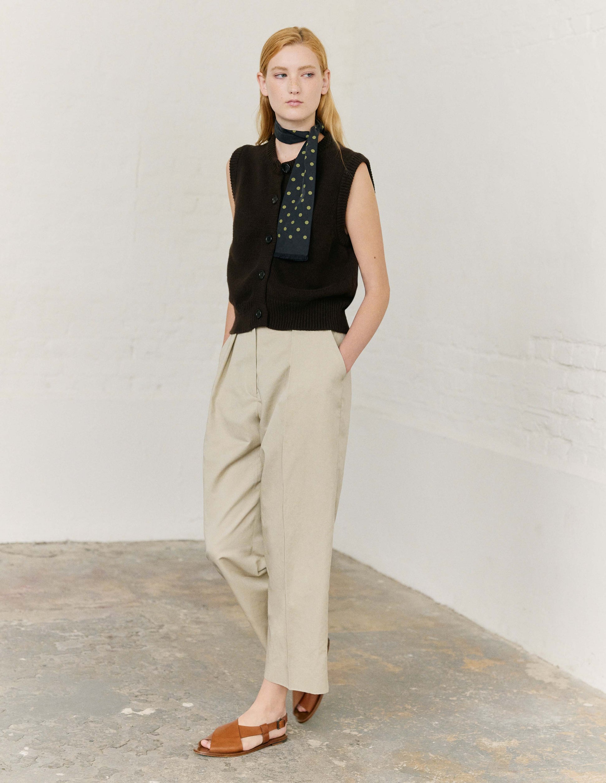 Full body image, front view of model wearing dark brown sleeveless merino cashmere gilet with contrast rib neckline, hem and dark horn buttons. Styled with beige trousers, dark navy blue and yello polka dot silk scarf, and light brown leather sandals.